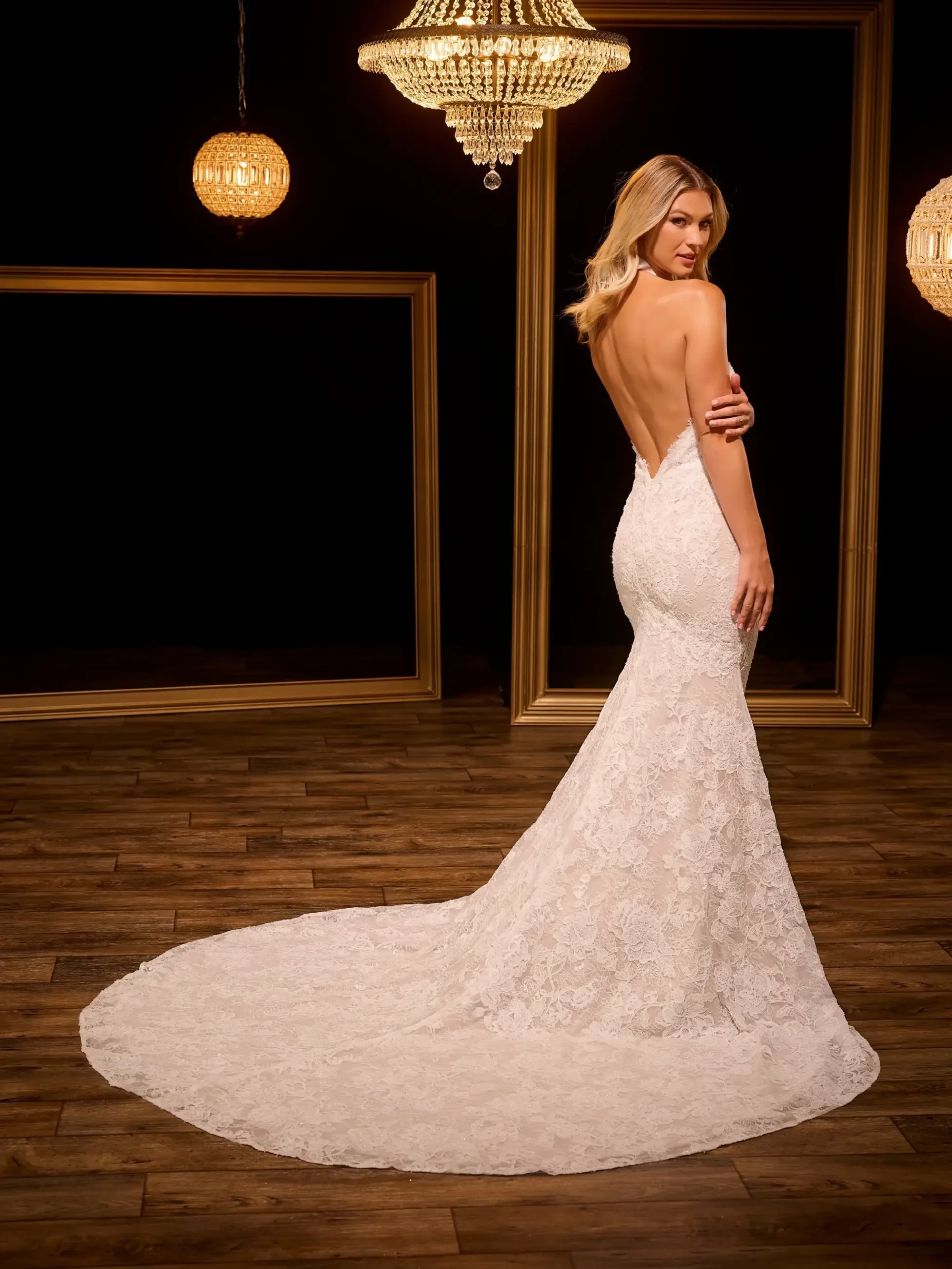A woman in a backless lace bridal gown stands on a wooden floor, her train gracefully spread. Golden chandeliers and frames create an elegant backdrop.