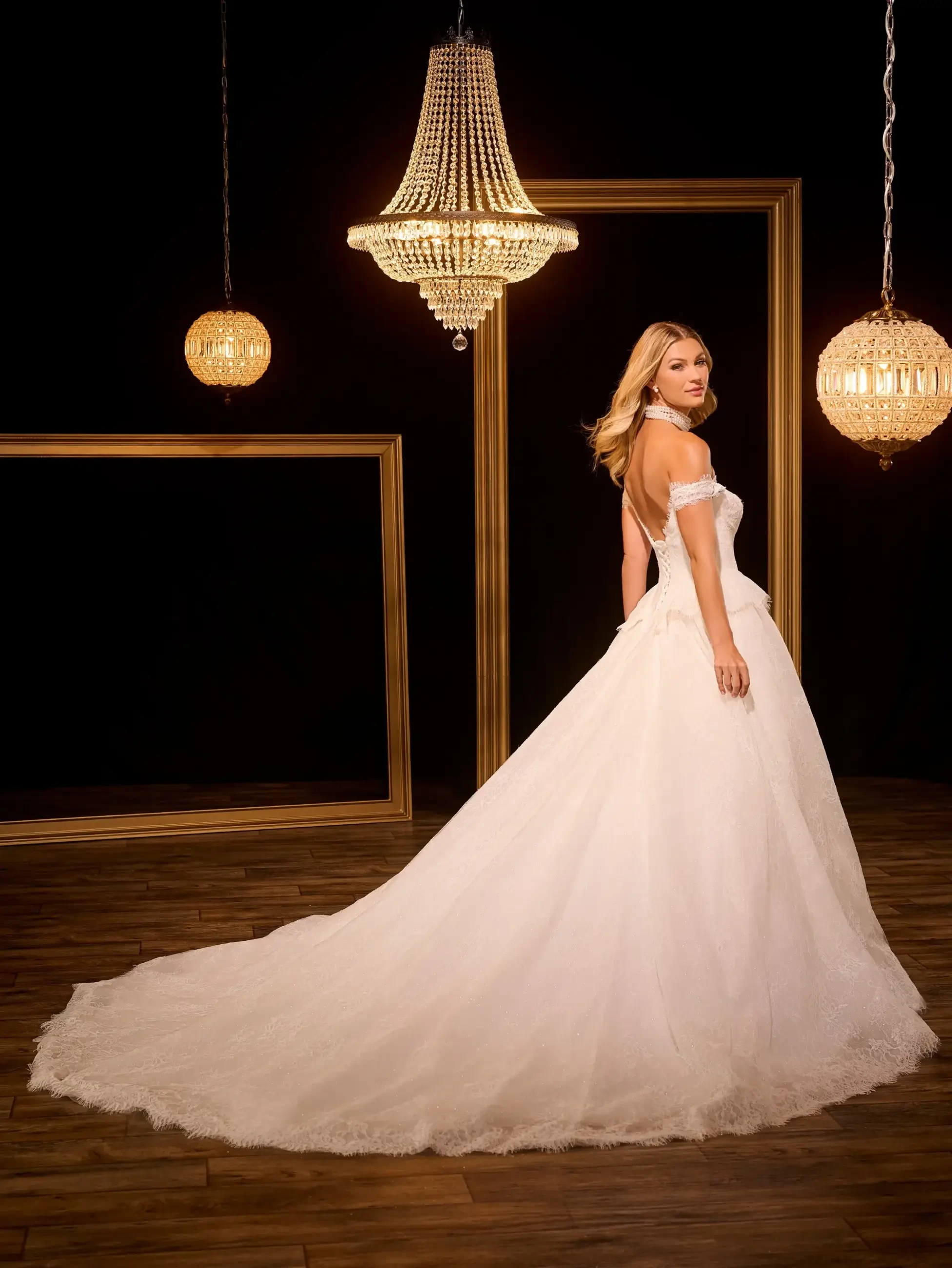 A woman in a white wedding gown with an off-the-shoulder design stands facing away from the camera, showcasing the back of her dress. The setting features elegant chandeliers and framed mirrors.