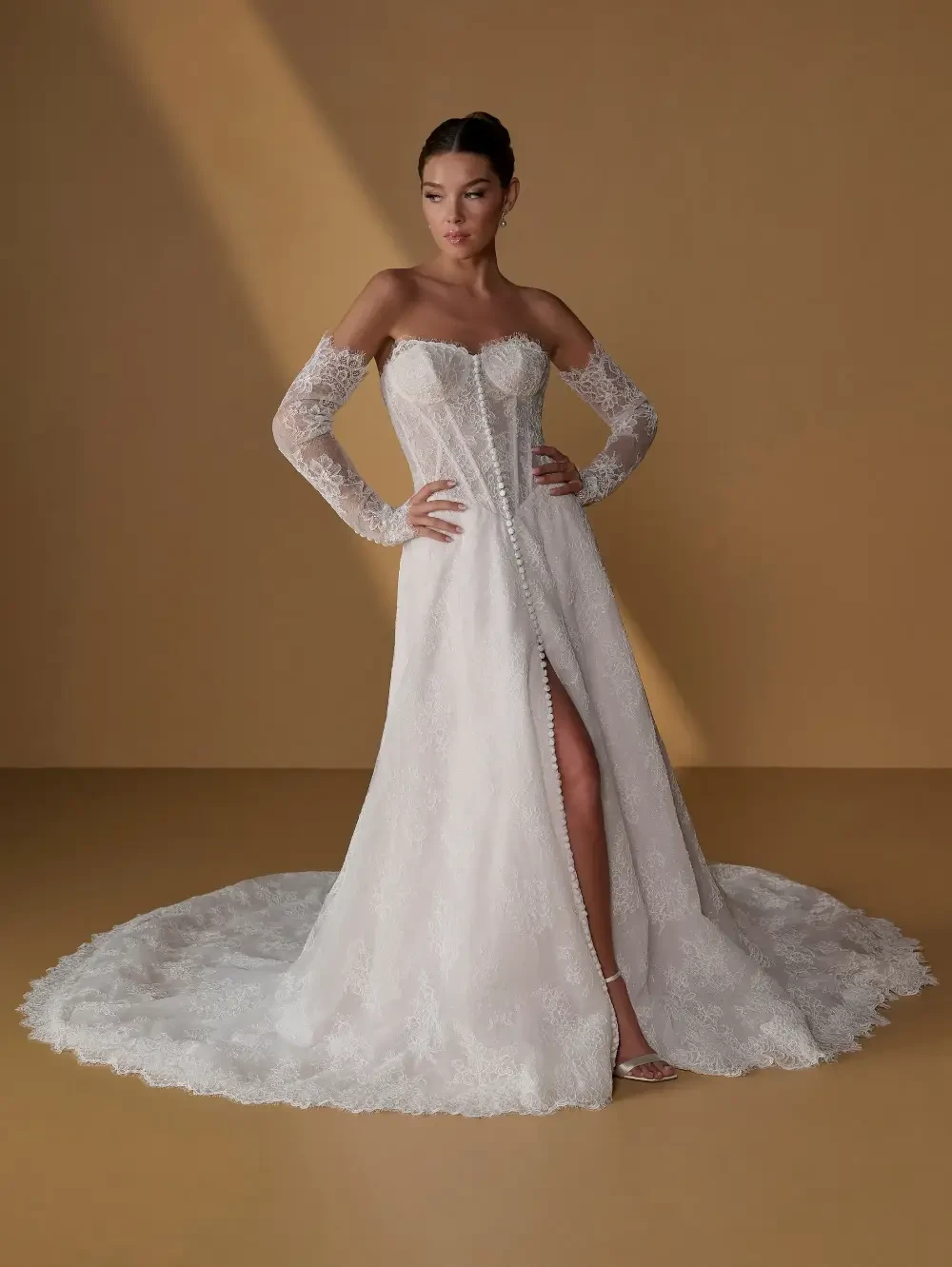A model wearing a strapless white wedding dress with lace details and long sheer sleeves, featuring a thigh-high slit and a train, posed against a warm-toned background.