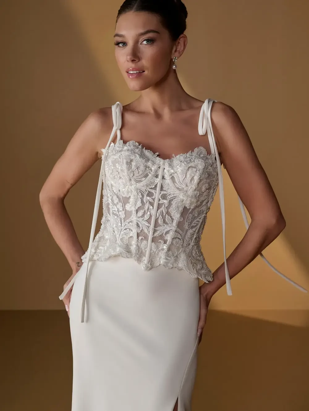 A woman wearing an elegant white dress with intricate beading and lace details on the bodice, posed in front of a warm-toned background.
