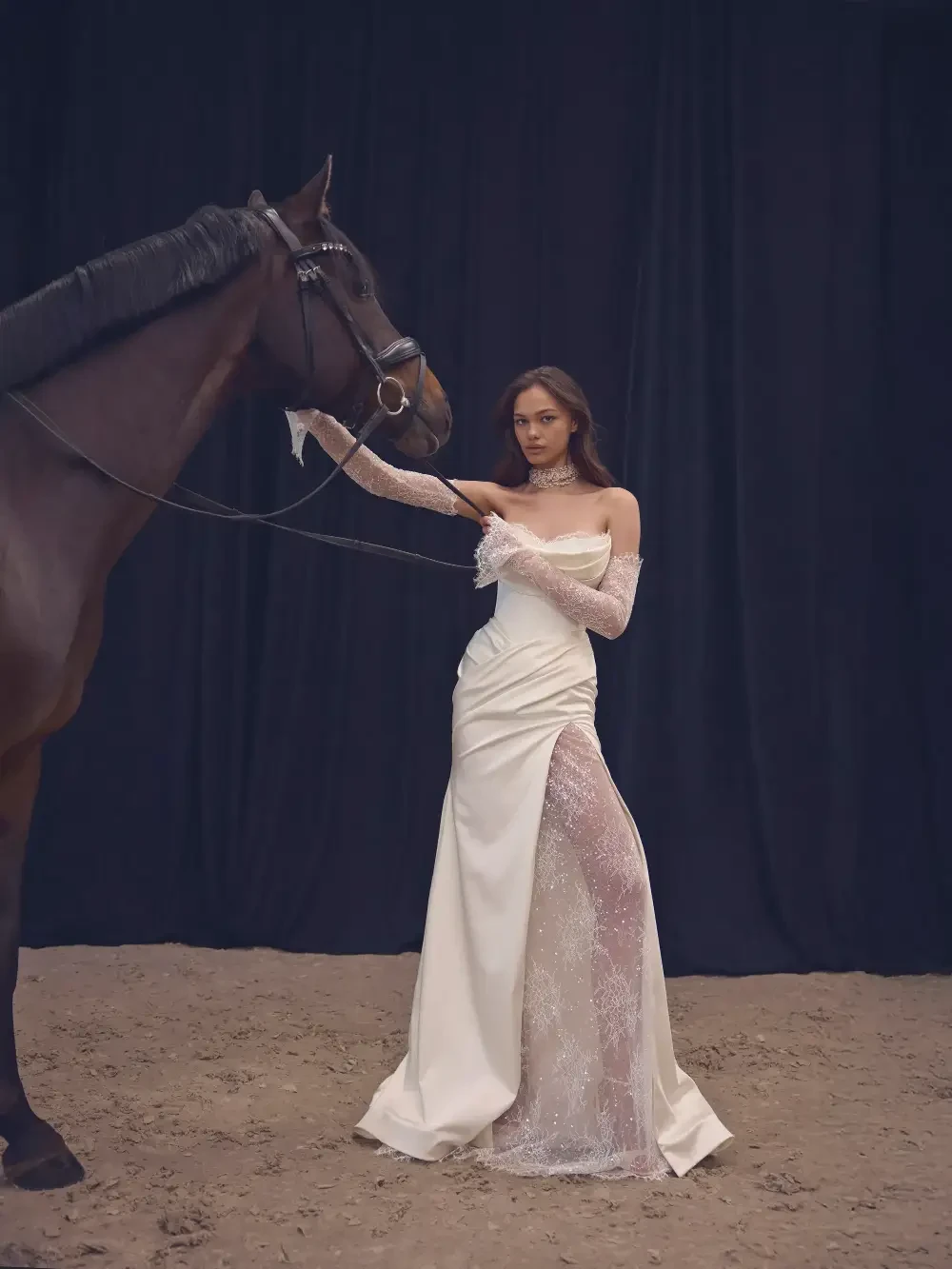 A woman in a white gown stands beside a horse against a dark backdrop.