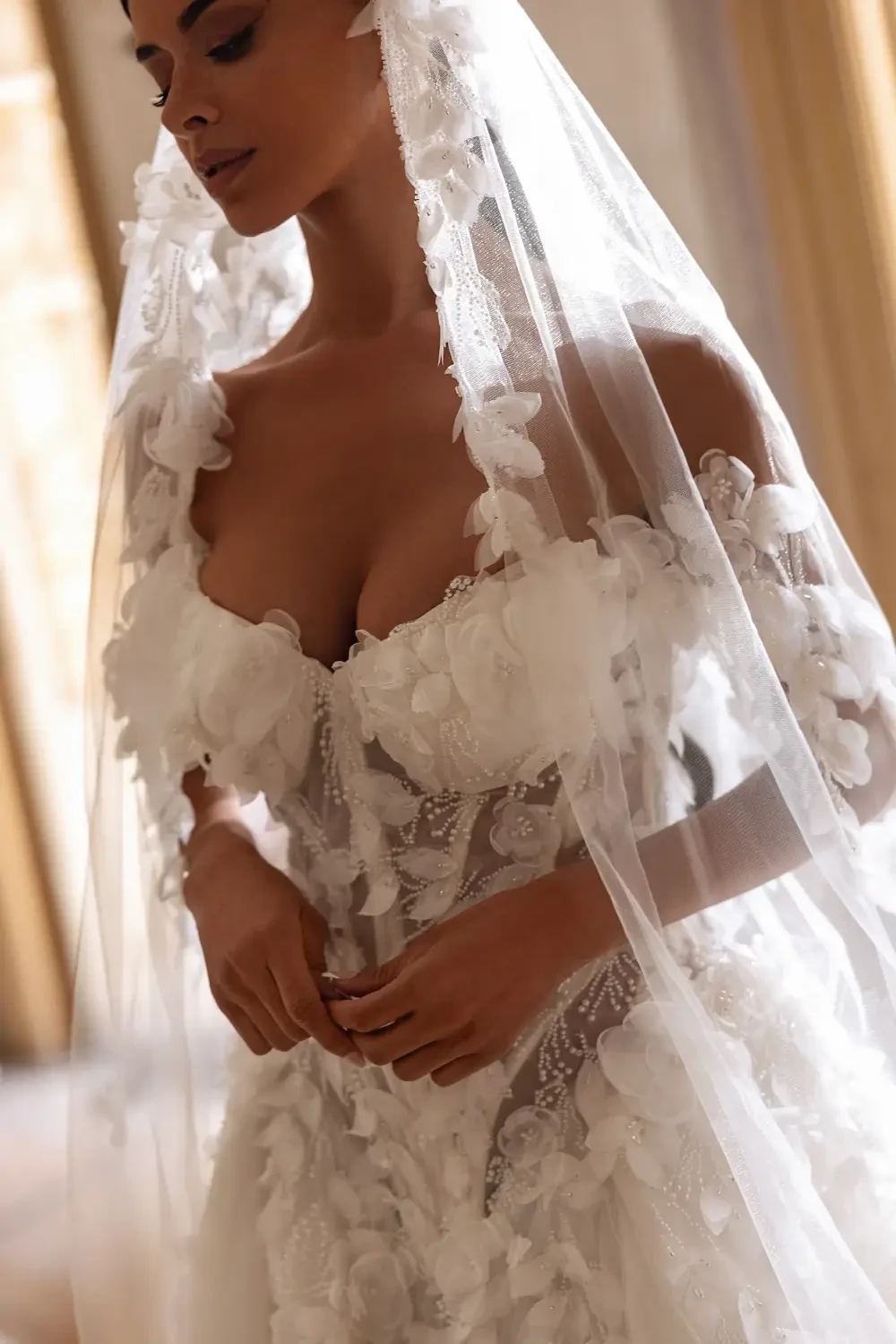 A bride in a detailed white wedding dress with lace and a long veil stands in a room with large windows and natural light.