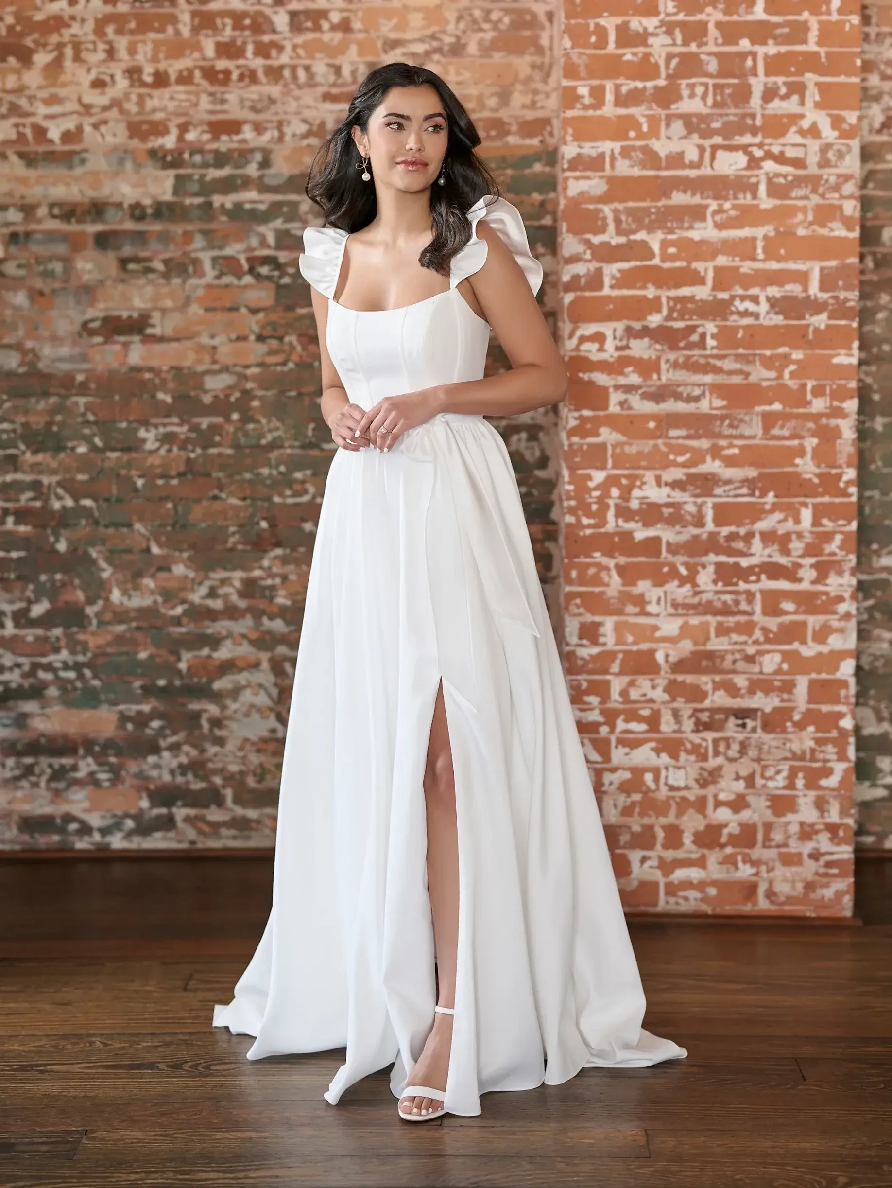 Model is wearing a satin gown with encased boning on the bodice, a self-tie belt, soft gathers in the skirt, and a ruffle sleeve.