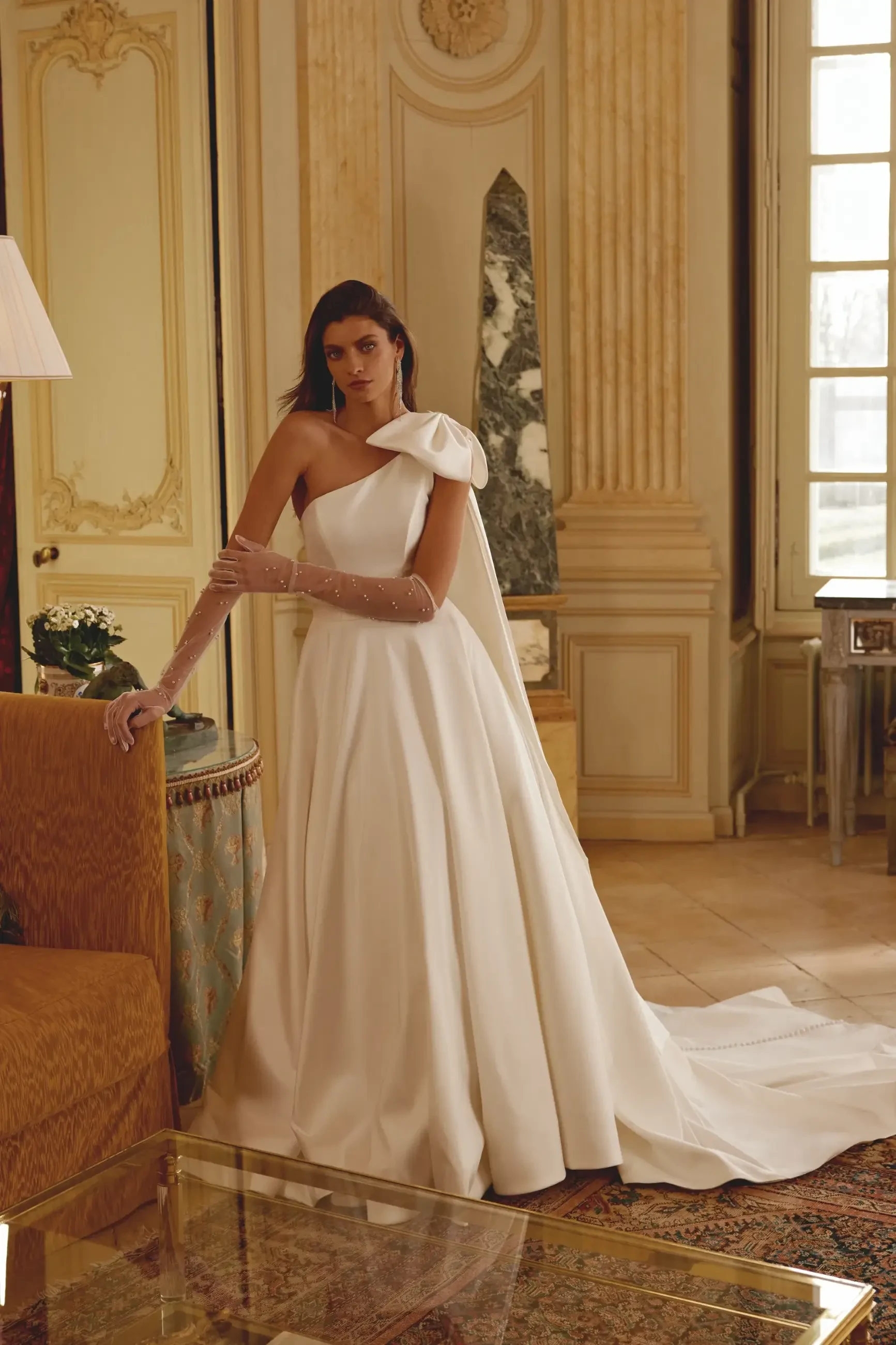 model is wearing a one shoulder wedding dress with a large bow