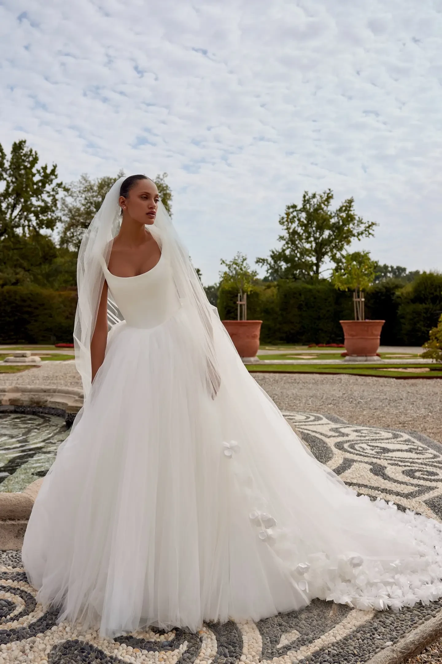 Model is wearing a tulle ball gown with a Basque waistline and delicate bow accents trailing down the back.