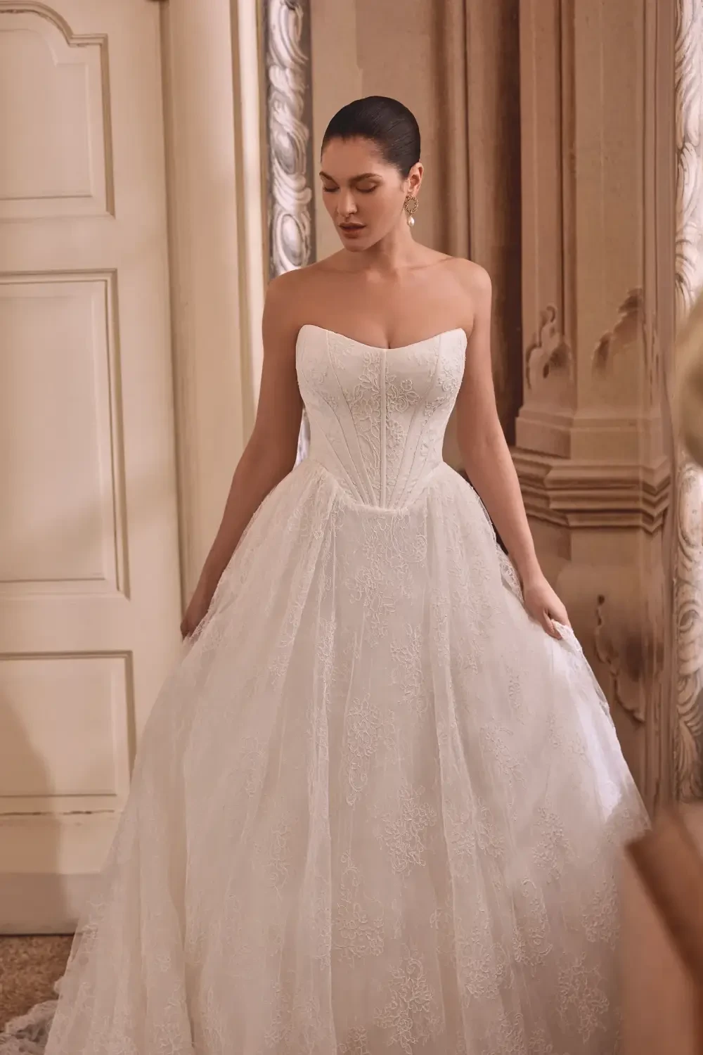 A woman wearing a strapless white wedding dress stands elegantly in an ornate room.