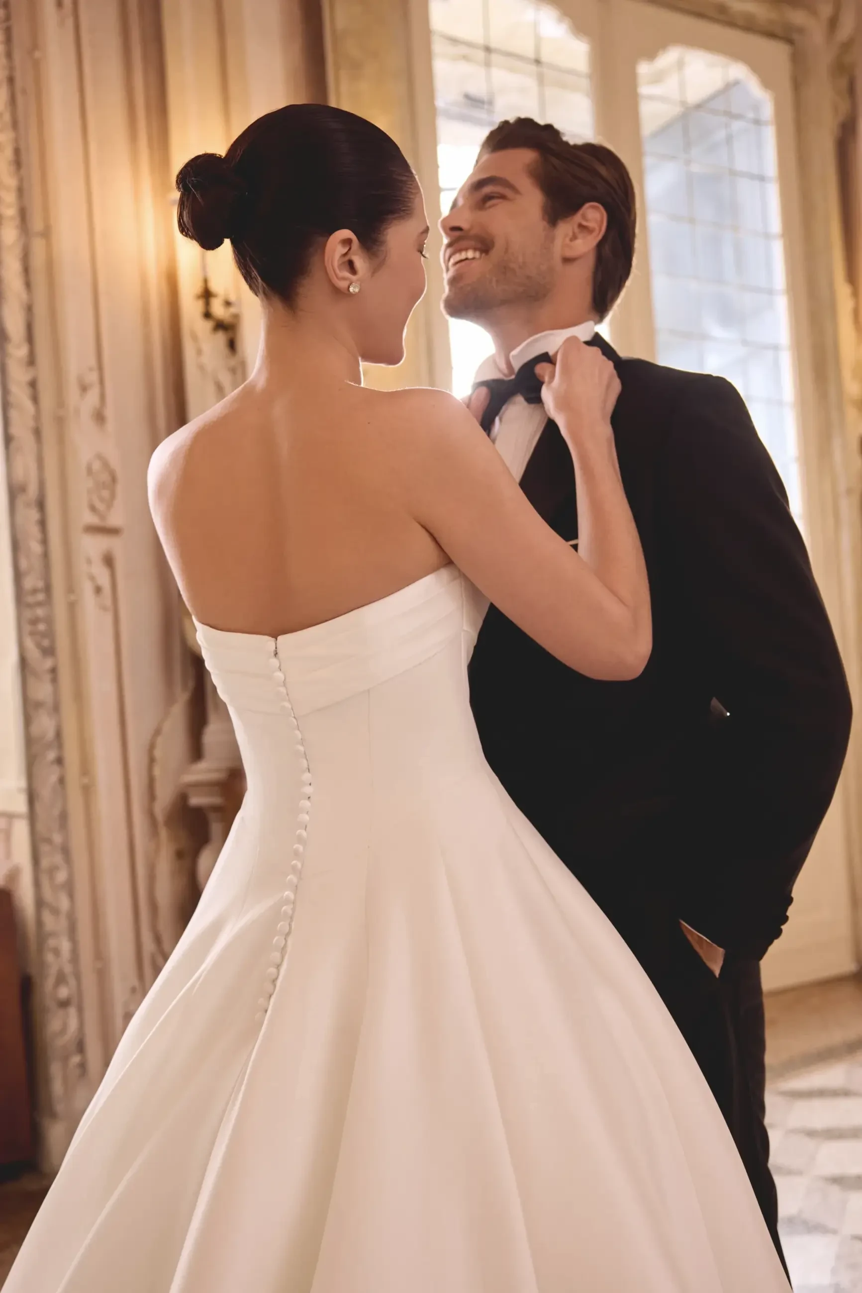 A couple standing close together, with the woman wearing a strapless white wedding gown and the man in a black tuxedo, sharing a moment in an elegant setting.