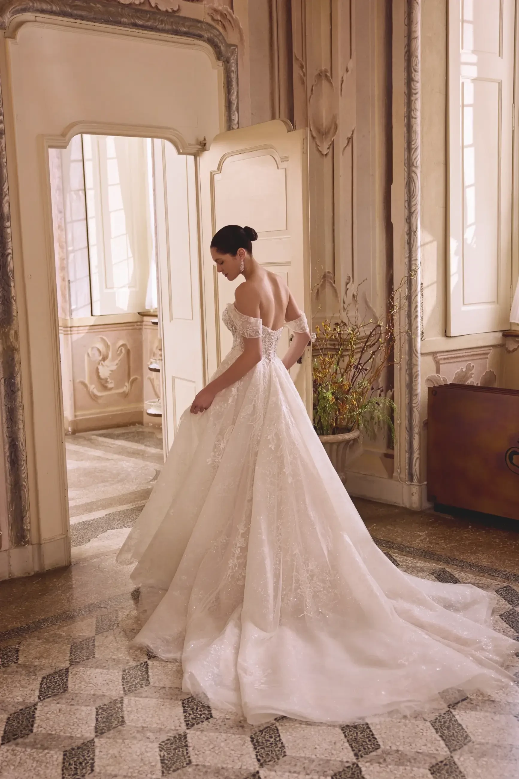 A bride in an elegant off-shoulder gown stands in an ornate room with vintage decor and sunlight streaming through large windows, creating a serene atmosphere.