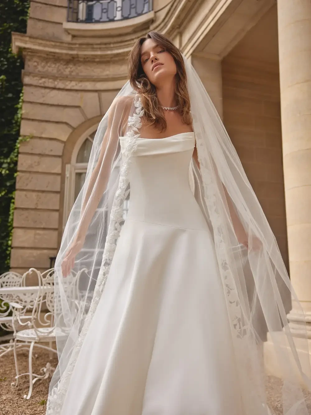 A model wearing a white wedding dress and a long veil, standing outdoors in a classic architectural setting.