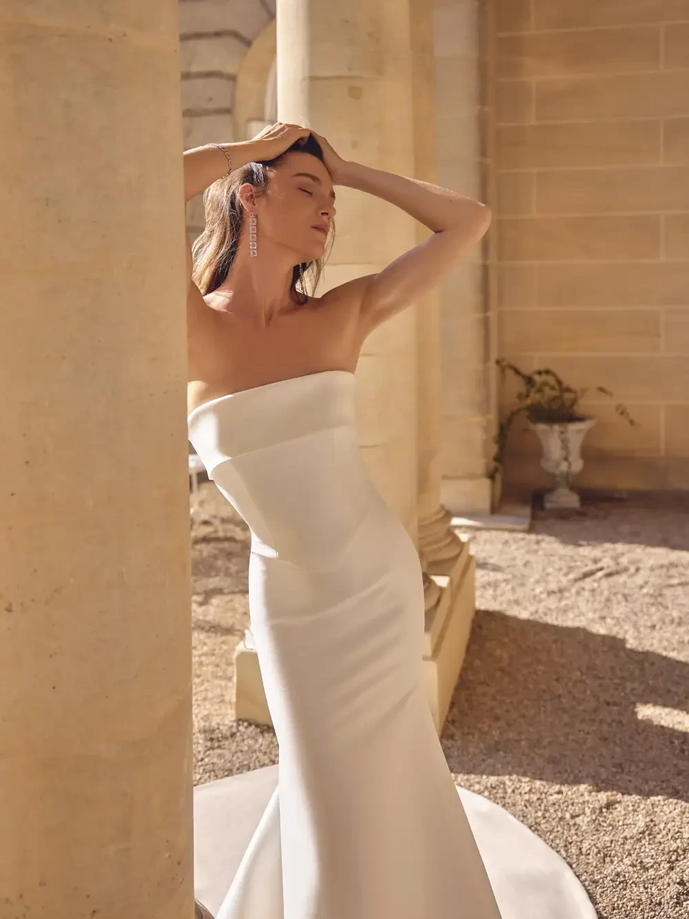 A woman in a strapless white gown posing elegantly near a column in a well-lit outdoor setting.