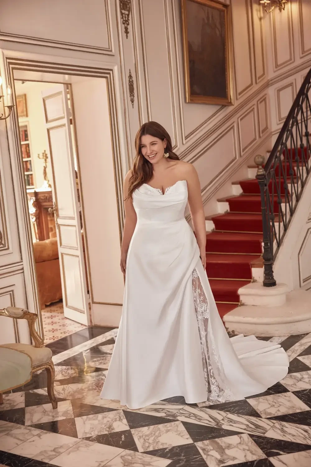 A woman stands in a beautiful white strapless wedding dress with a slit and lace detail, smiling in an elegant interior with marble flooring and a staircase.