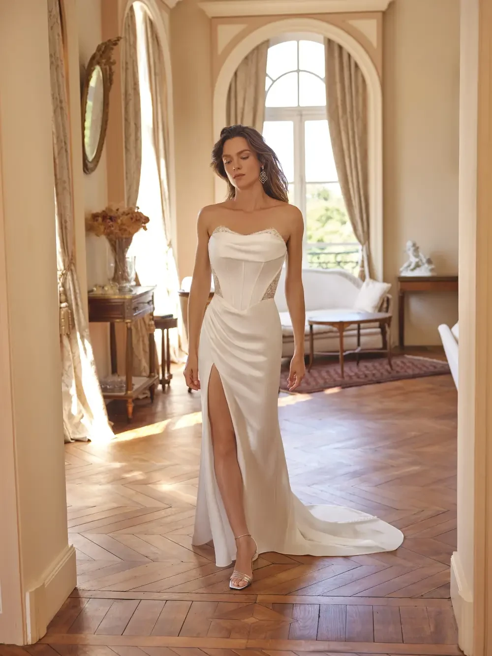 A woman in a strapless white dress with a thigh-high slit standing in a beautifully decorated room with large windows.