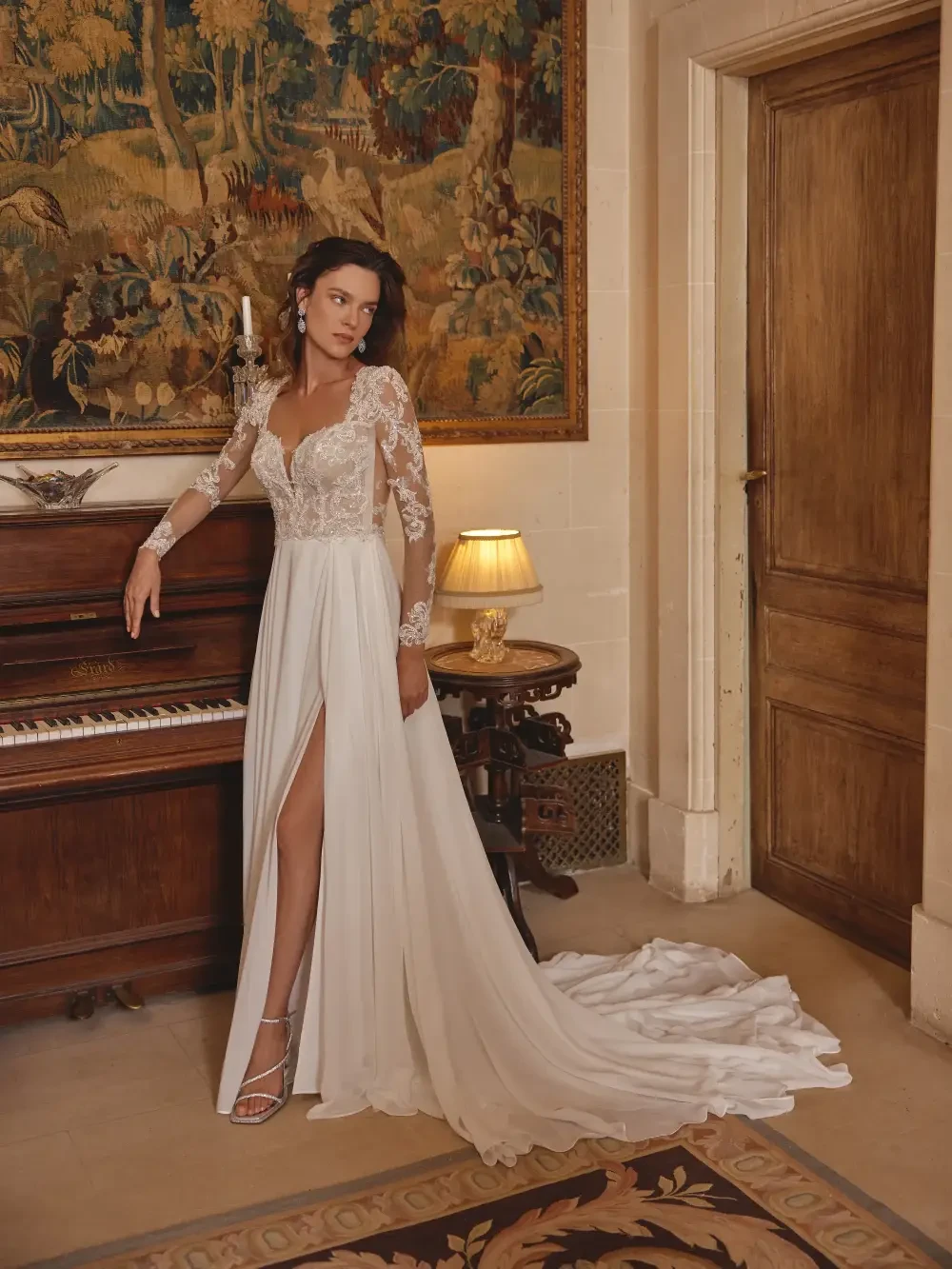A woman in a white wedding gown with long lace sleeves and a high slit stands next to a piano in an elegant room.