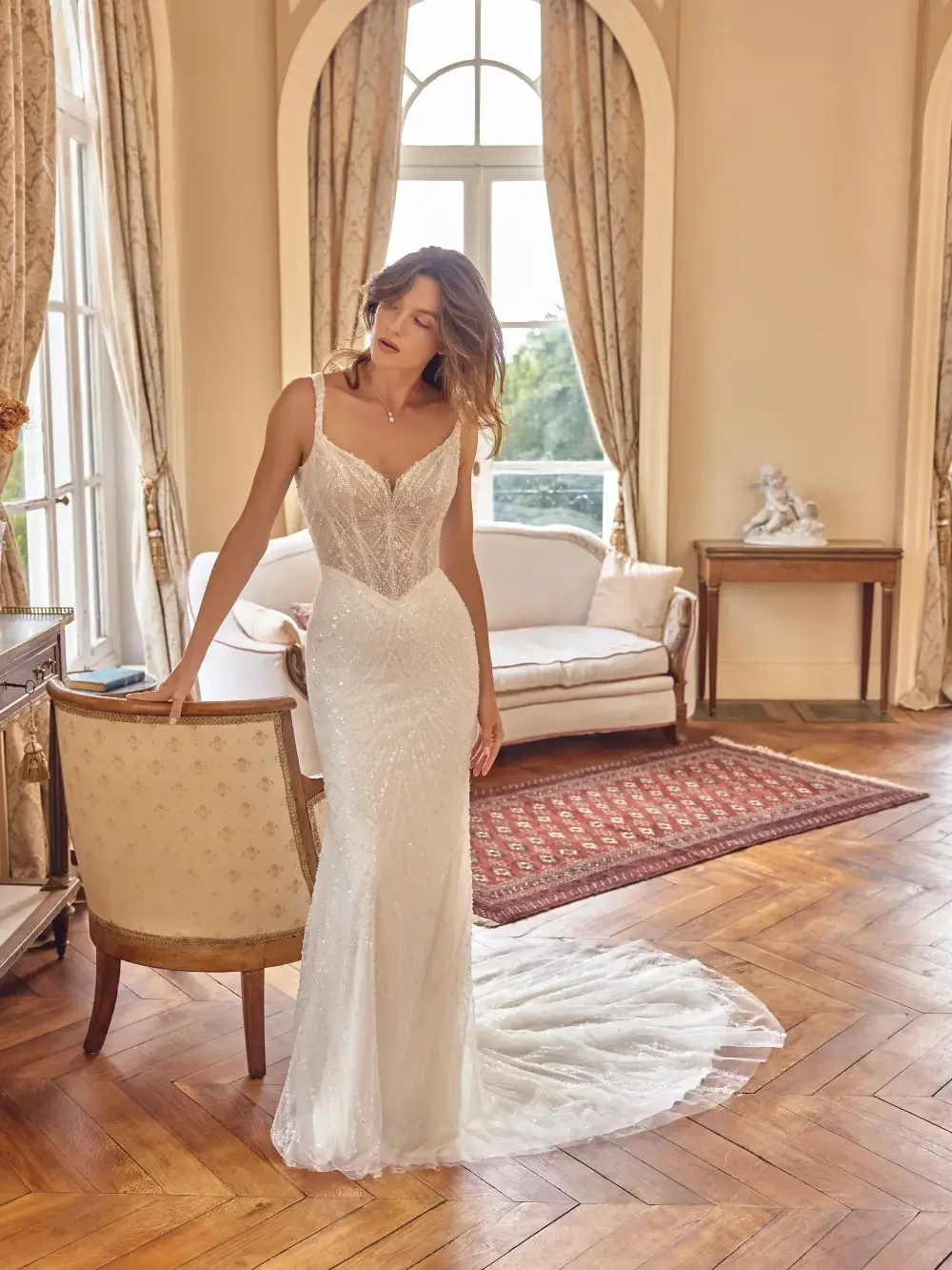 A woman in a sparkling white gown stands beside a chair in an elegantly decorated room with large windows and a plush sofa.