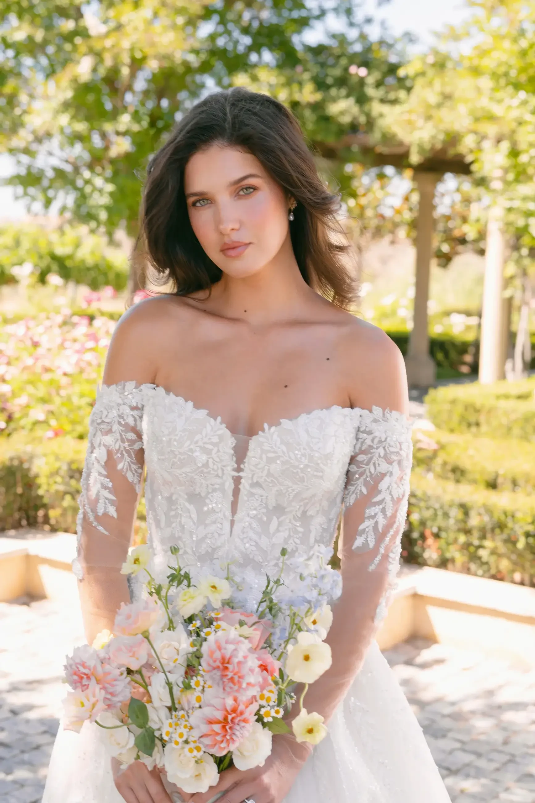 A woman in an elegant off-the-shoulder wedding dress holds a bouquet of flowers while standing in a garden.