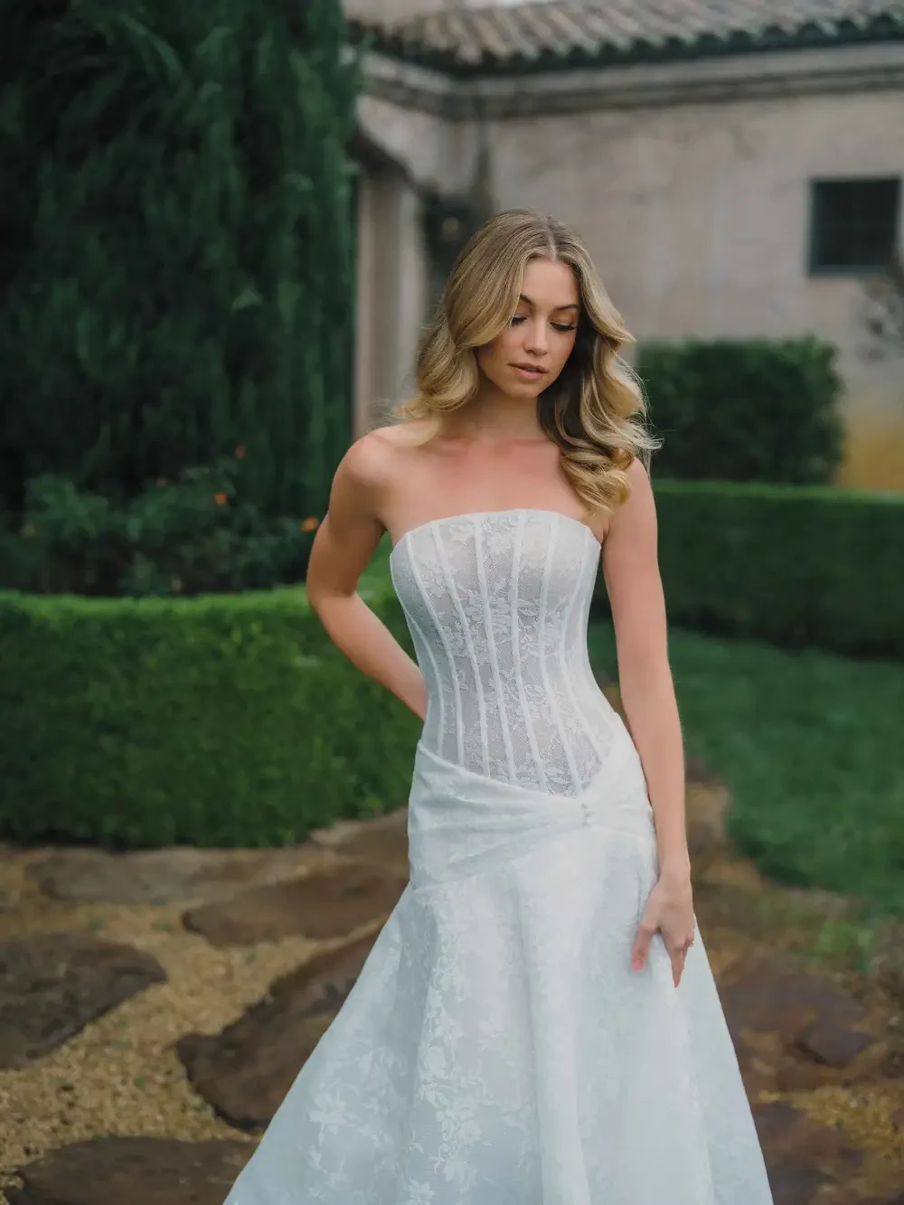 A woman in a strapless wedding dress stands gracefully outdoors, with lush greenery and a stone pathway in the background.