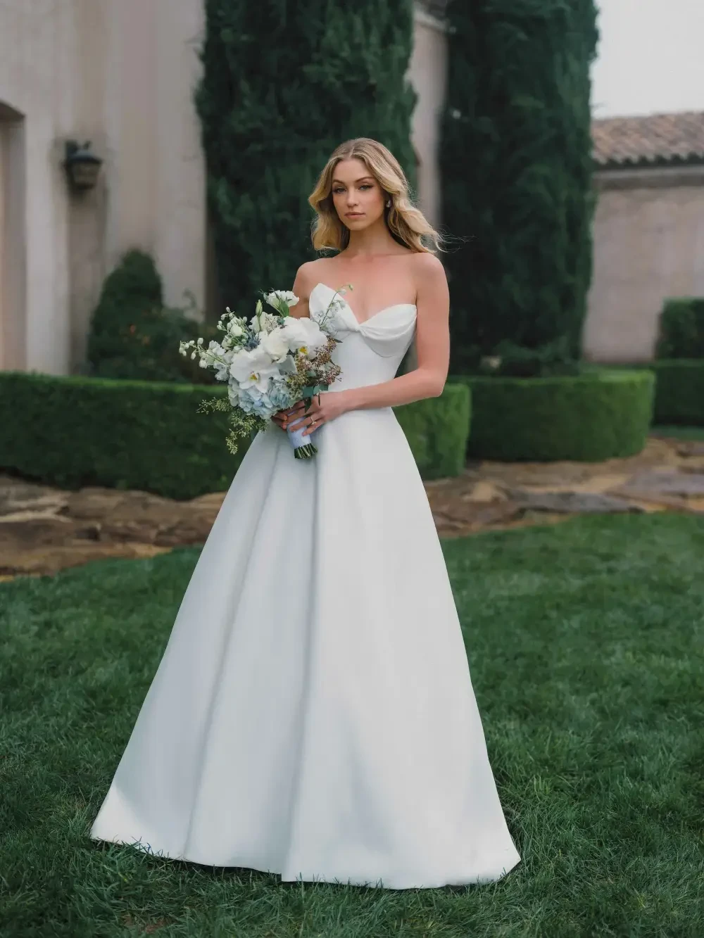 A woman in a strapless white wedding gown holding a bouquet of flowers, standing outside on a grassy area with hedges and a building in the background.