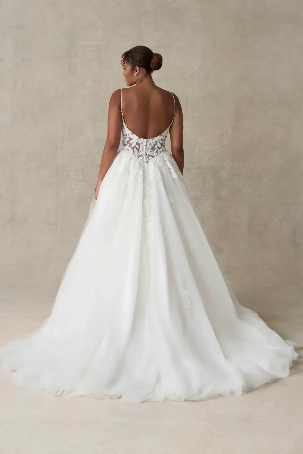 A woman wearing a backless white wedding dress with intricate beading, standing against a neutral backdrop.
