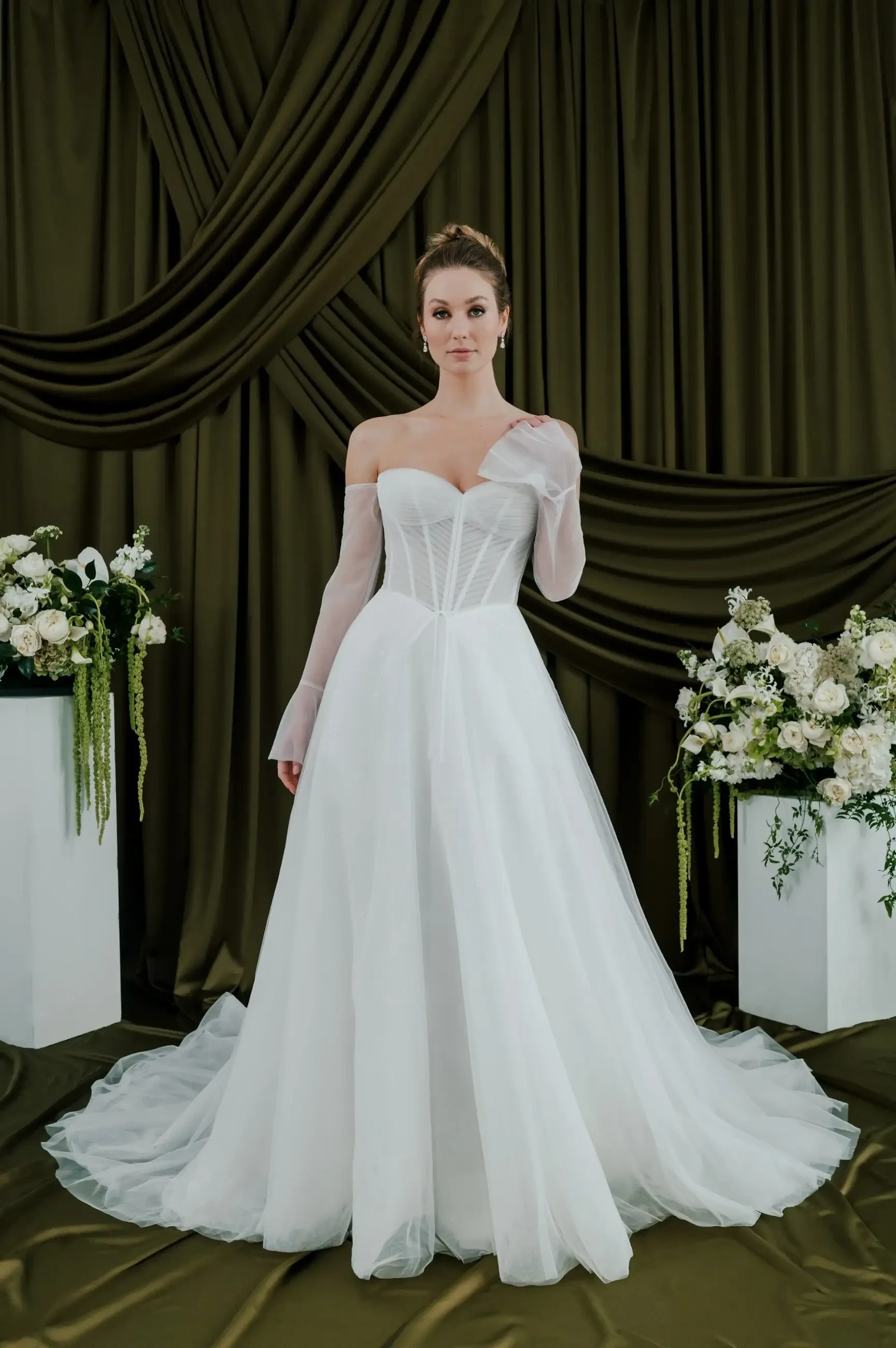 Bride in elegant white gown with sheer sleeves stands before draped olive-green curtains. Flanked by floral arrangements in white vases, exuding a serene and sophisticated vibe.