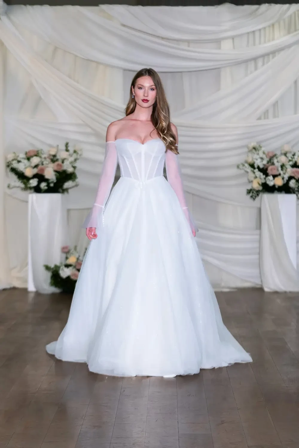 A model wearing a white off-the-shoulder wedding dress with sheer sleeves, standing on a wooden floor in front of floral arrangements and draped fabric.