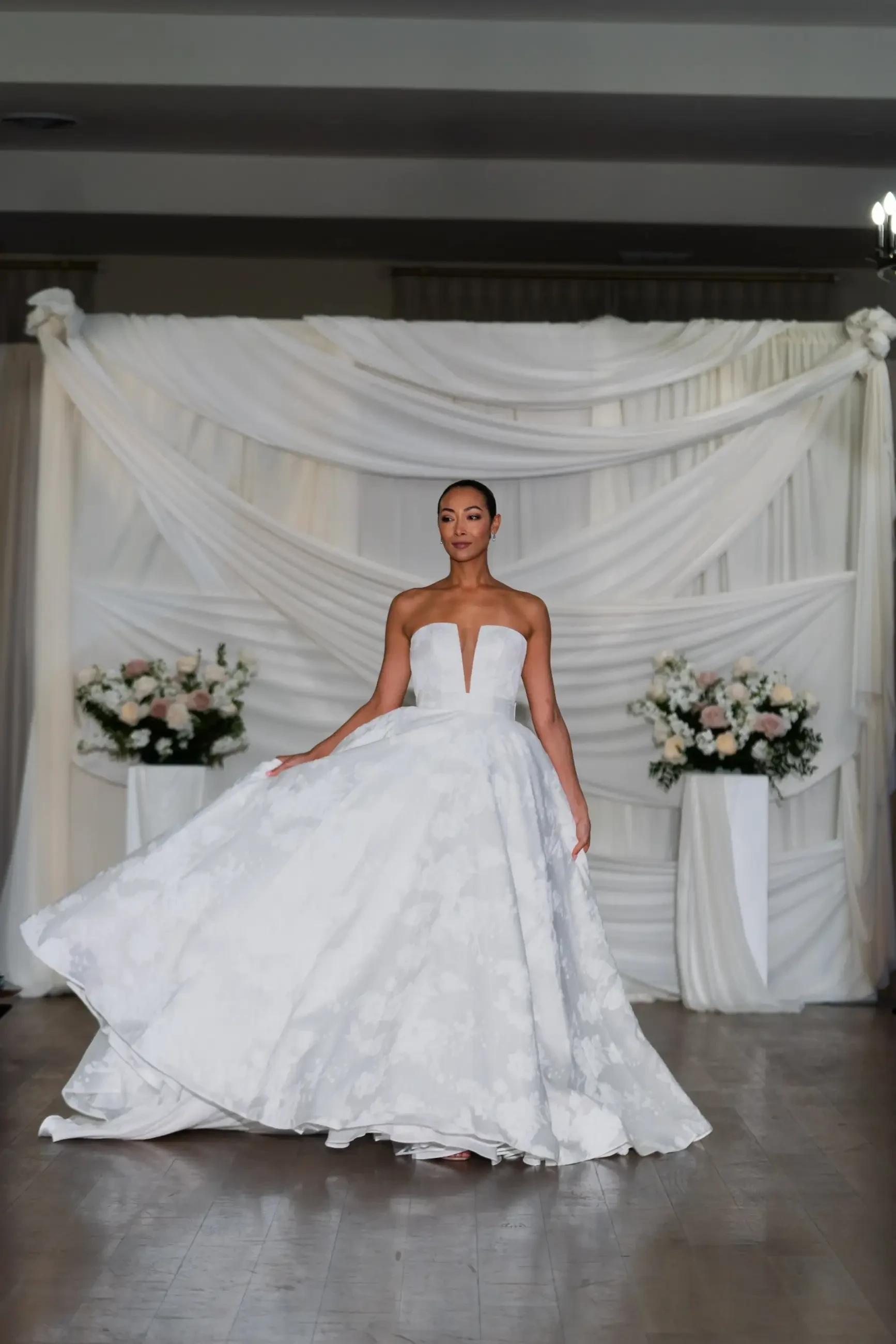 A model walks down a runway in a flowing white gown with a fitted bodice and no sleeves, surrounded by floral arrangements.