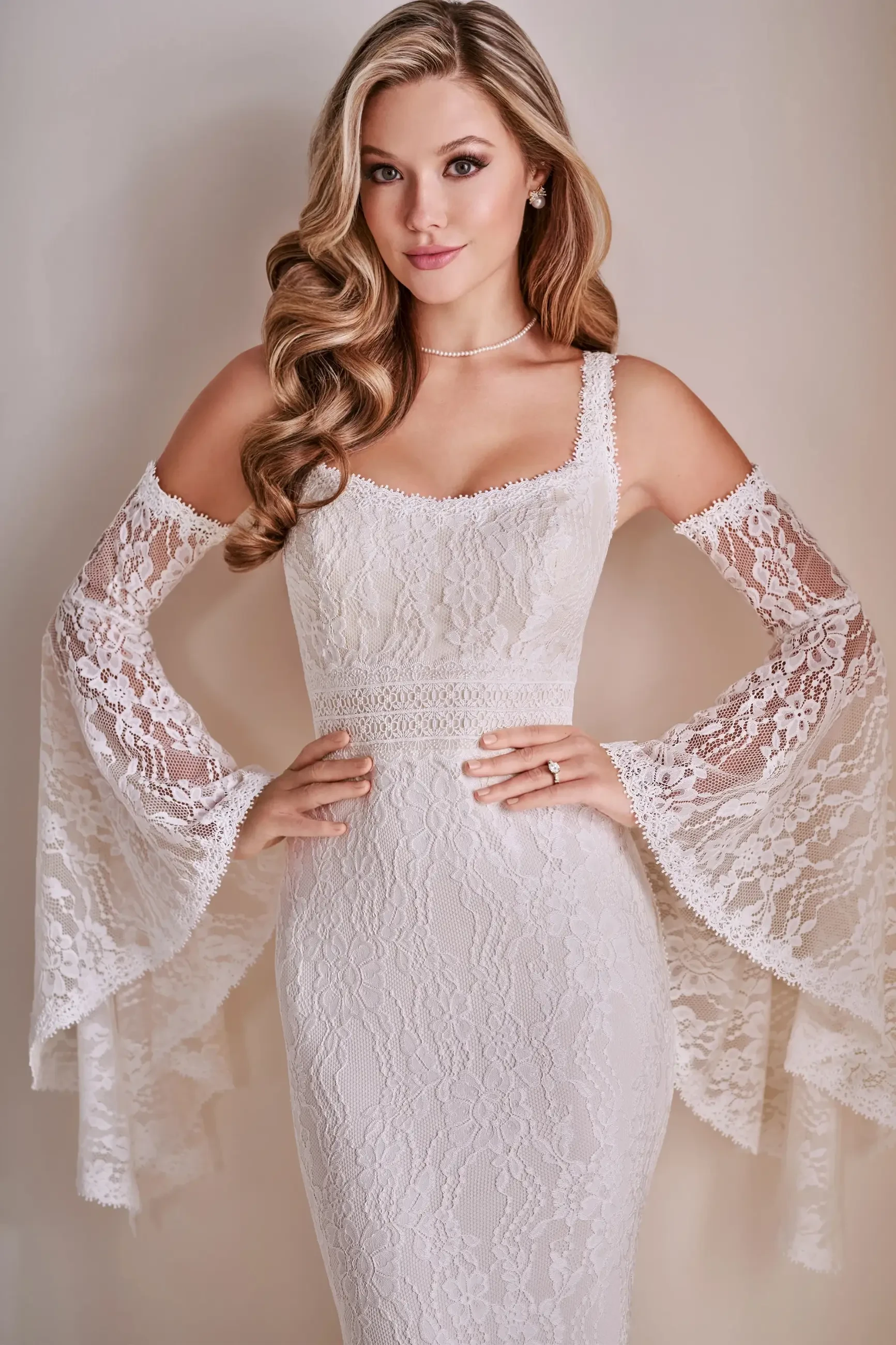 A woman wearing a white lace wedding dress with off-the-shoulder sleeves and loose lace overlays, styled with wavy hair and minimal jewelry.