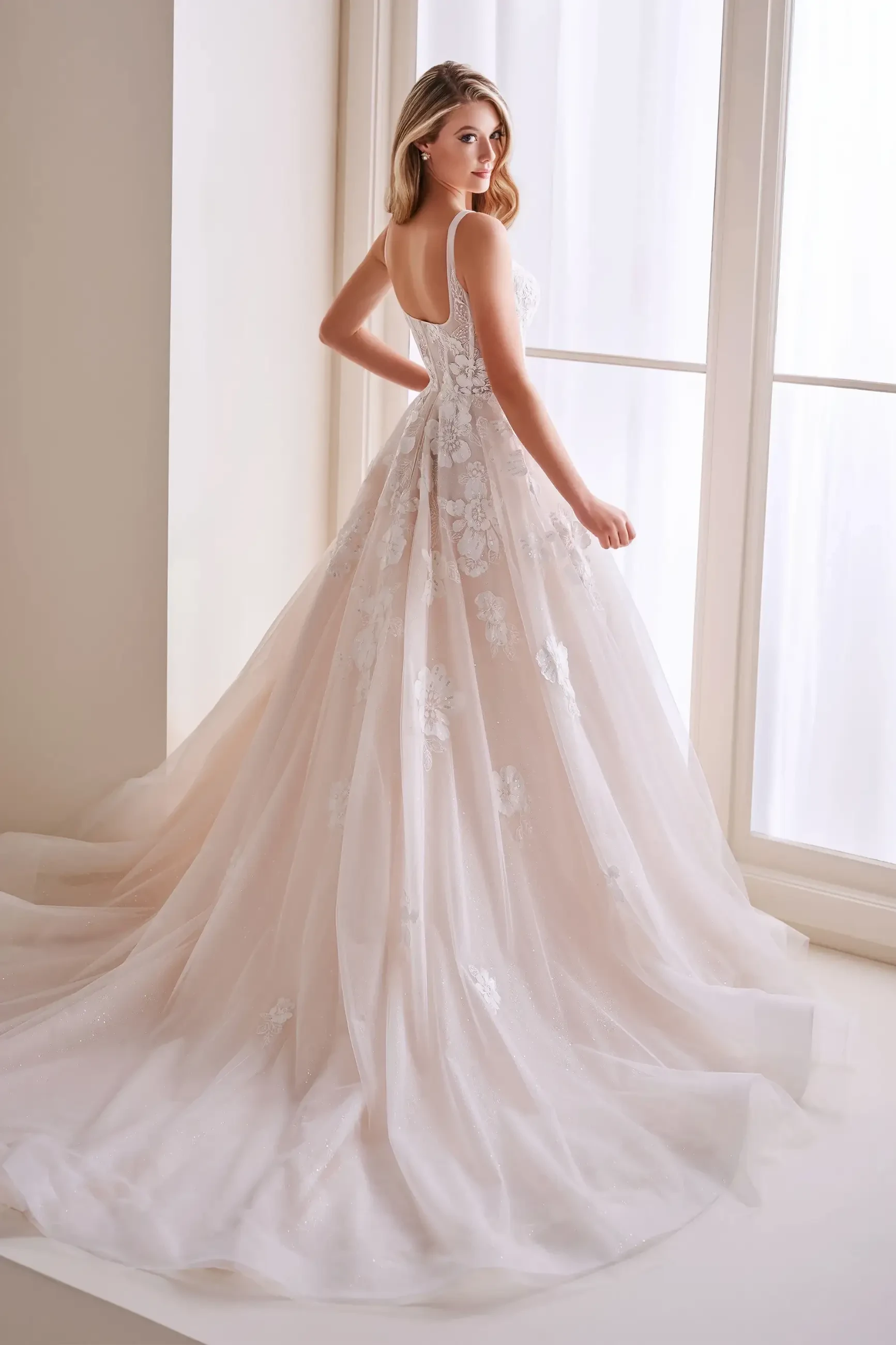 A woman wearing a beautiful wedding dress with intricate floral details, standing by a large window. The dress has a flowing skirt and a fitted bodice.