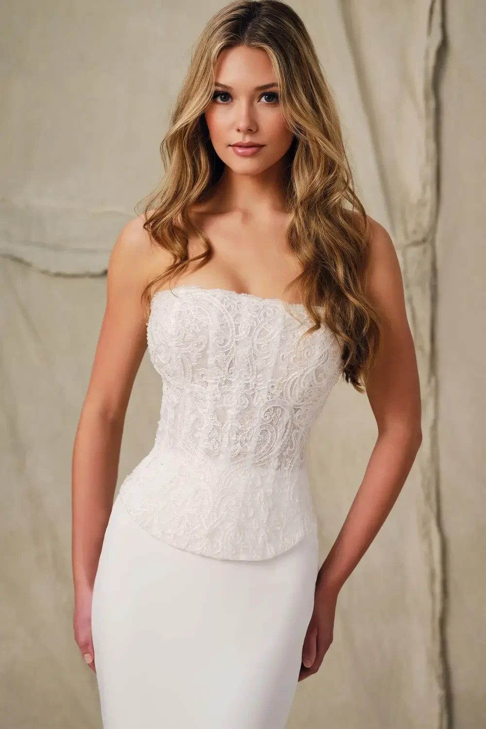 A woman poses in a strapless white dress with lace detailing at the bodice. She has long, wavy hair and a neutral expression against a soft, textured background.