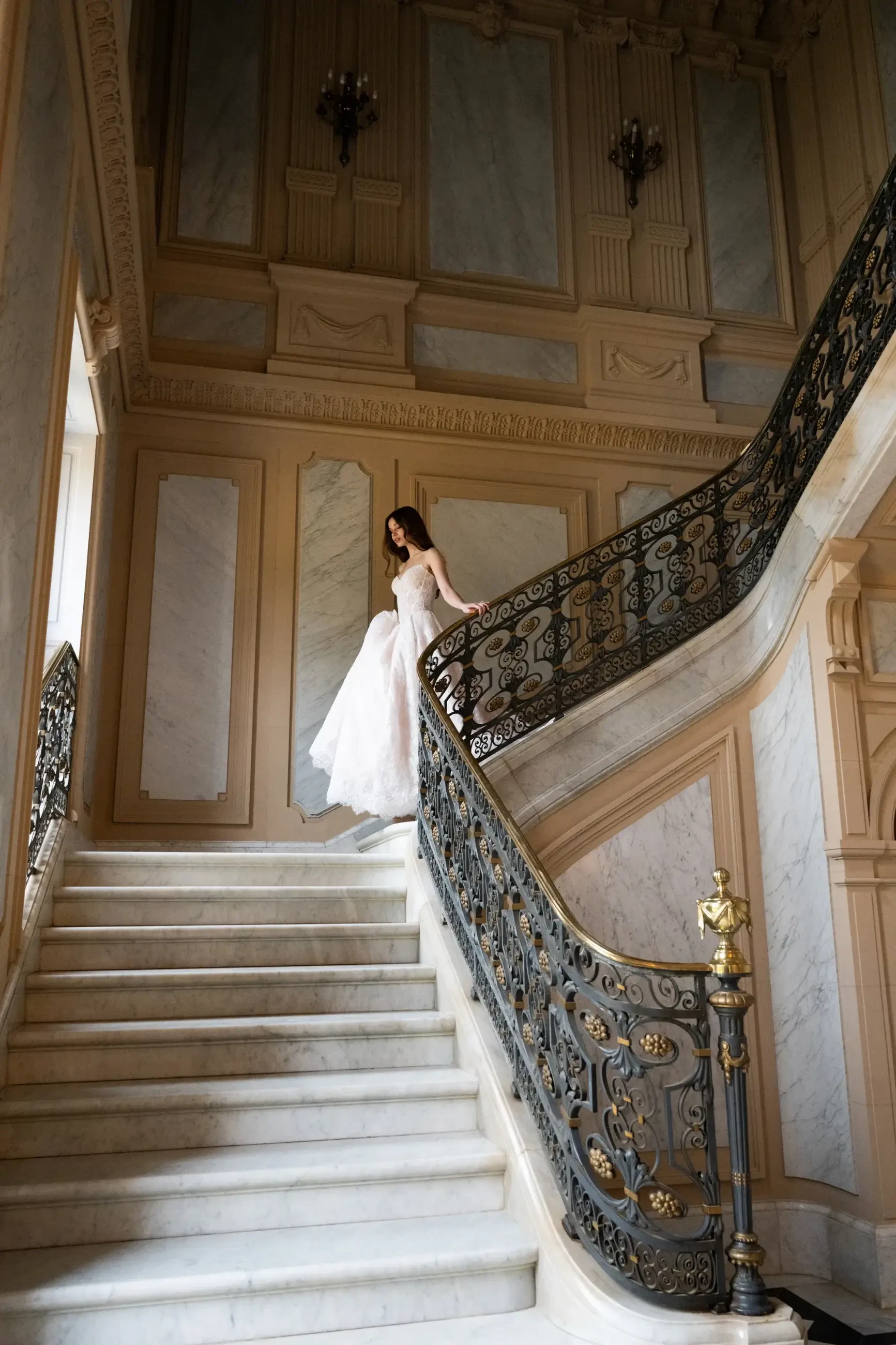 A woman in a wedding dress is descending a grand staircase with ornate iron railings and decorative walls.