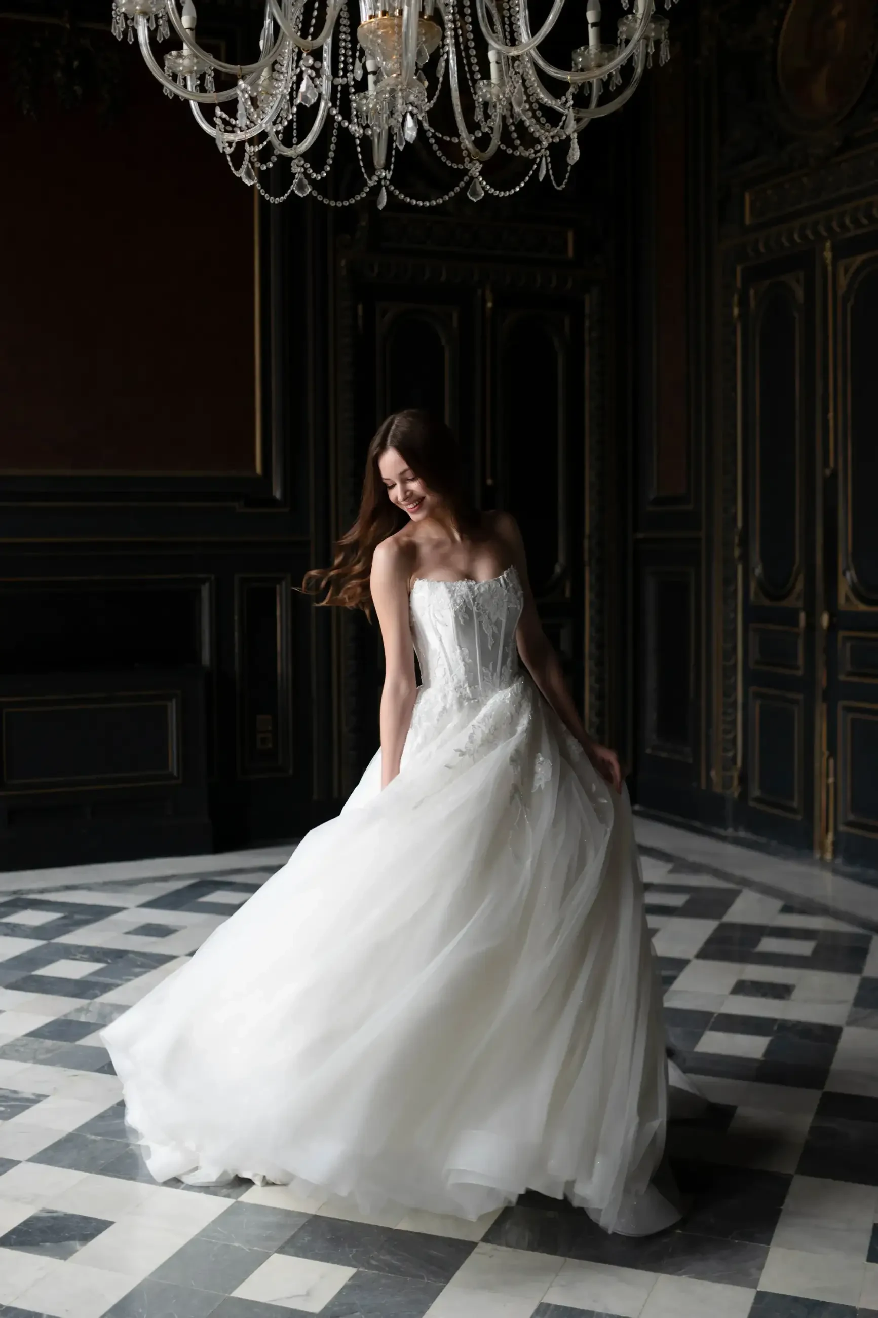 A woman in a flowing white wedding dress gracefully spins under an ornate chandelier in an elegant, dark-paneled room. She looks joyful and serene.