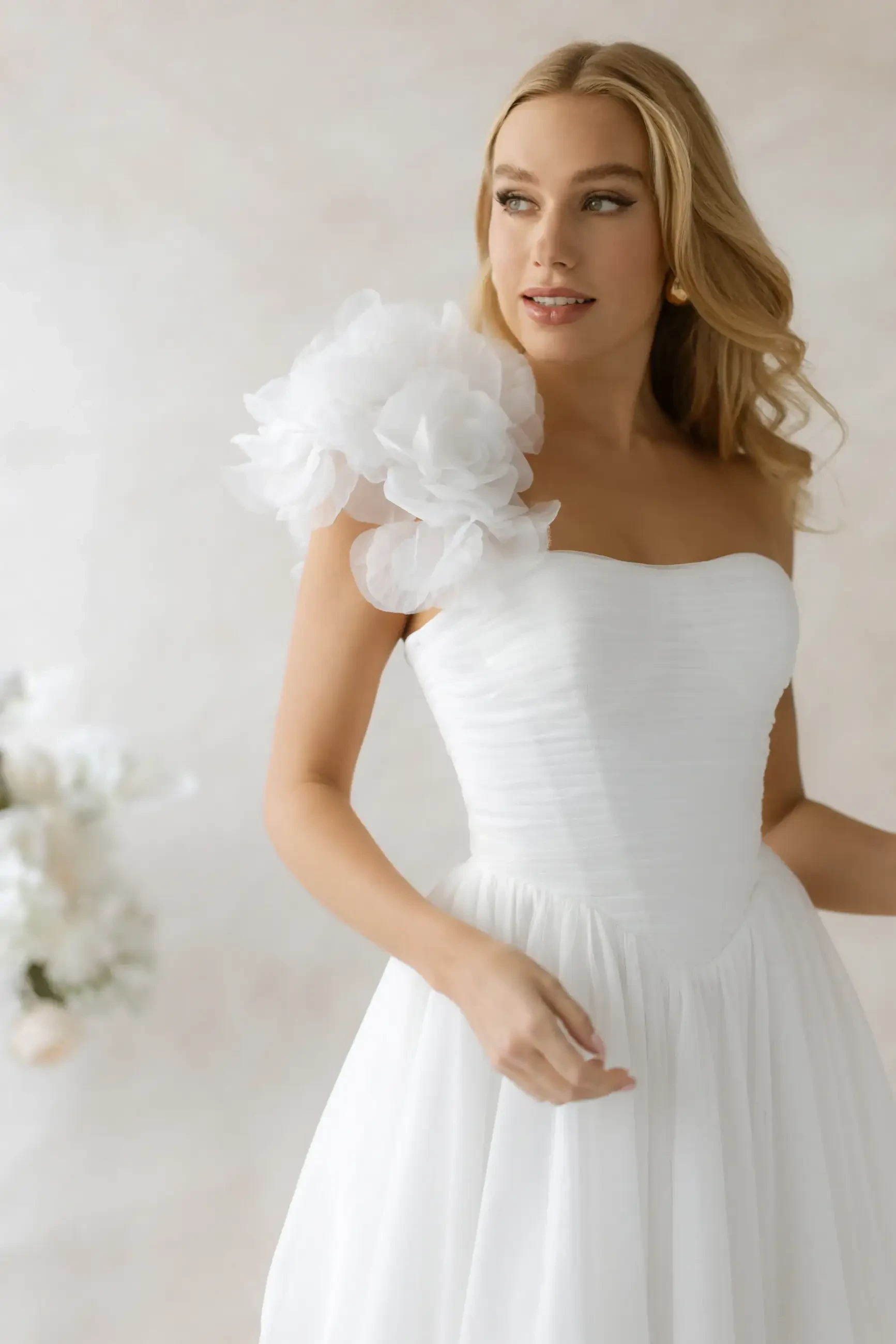 model is wearing a strapless bridal gown with a large floral detail on one shoulder