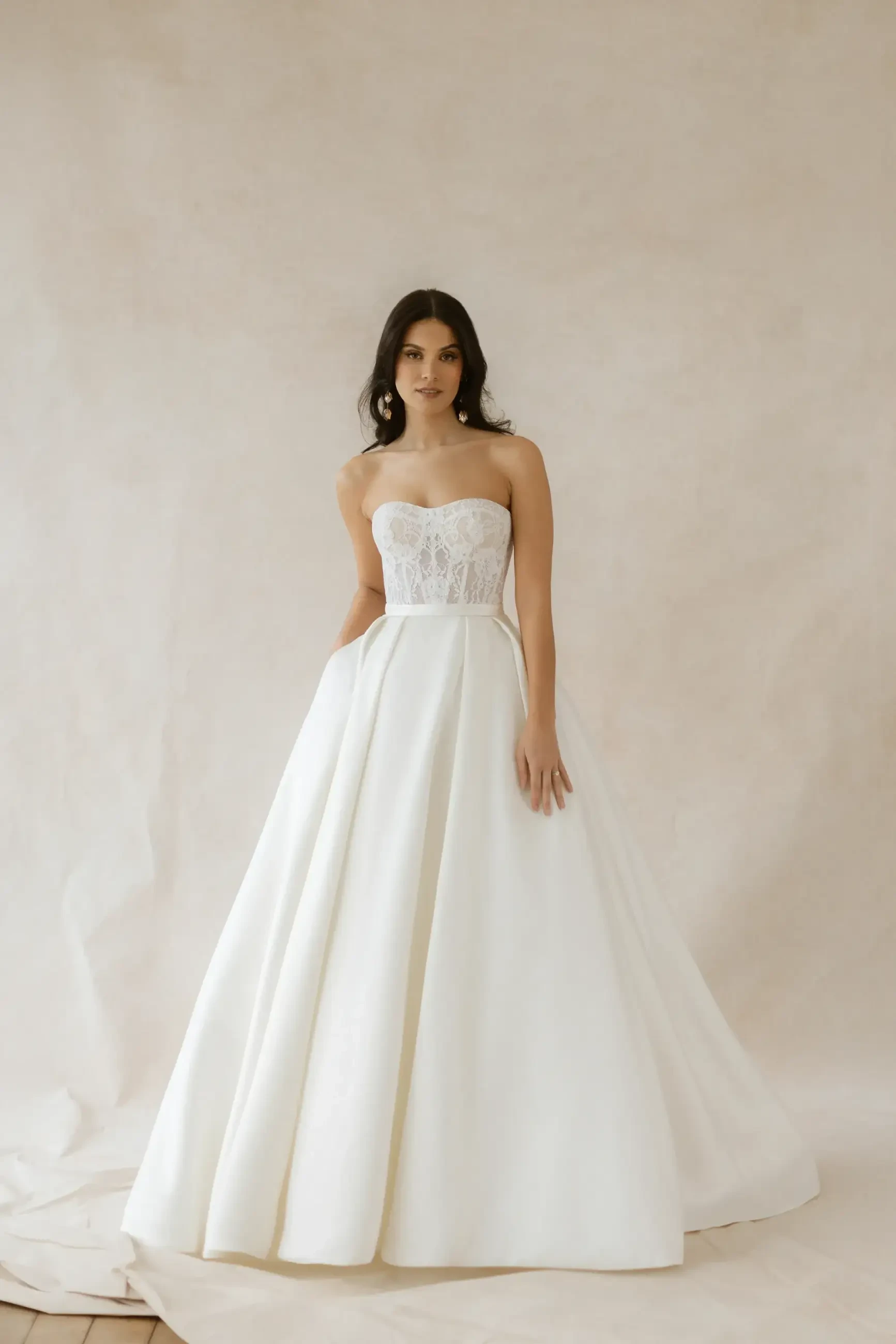 A woman in an elegant, strapless white wedding dress with a lace bodice and full skirt stands against a soft, neutral background, conveying grace.