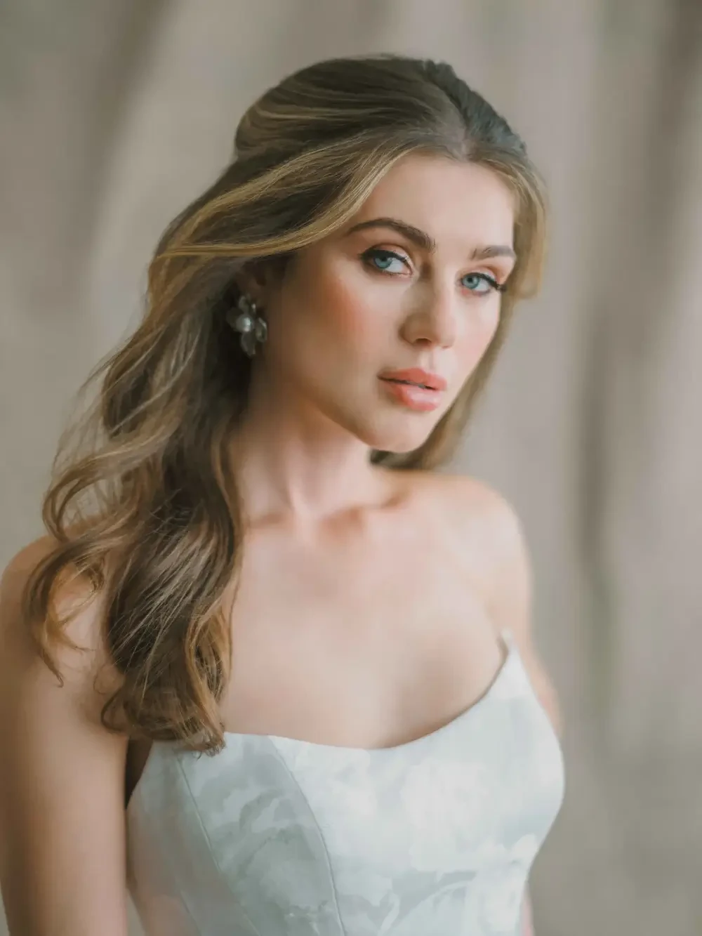 A woman with long, wavy hair and makeup wearing a white dress in a softly lit setting.