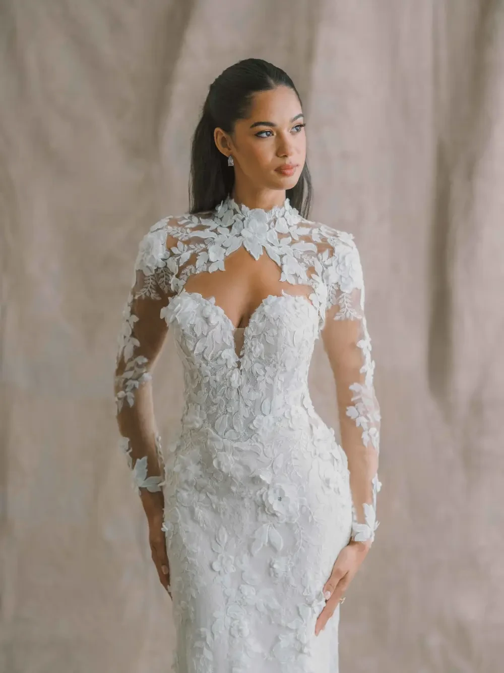 A woman in an elegant wedding dress with intricate floral details, featuring long sheer sleeves and a plunging neckline, posing against a textured background.