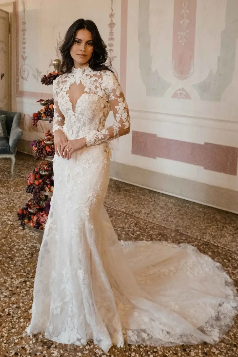 A woman wearing an elegant white lace wedding dress with long sleeves, posing in a vintage setting with floral decor.