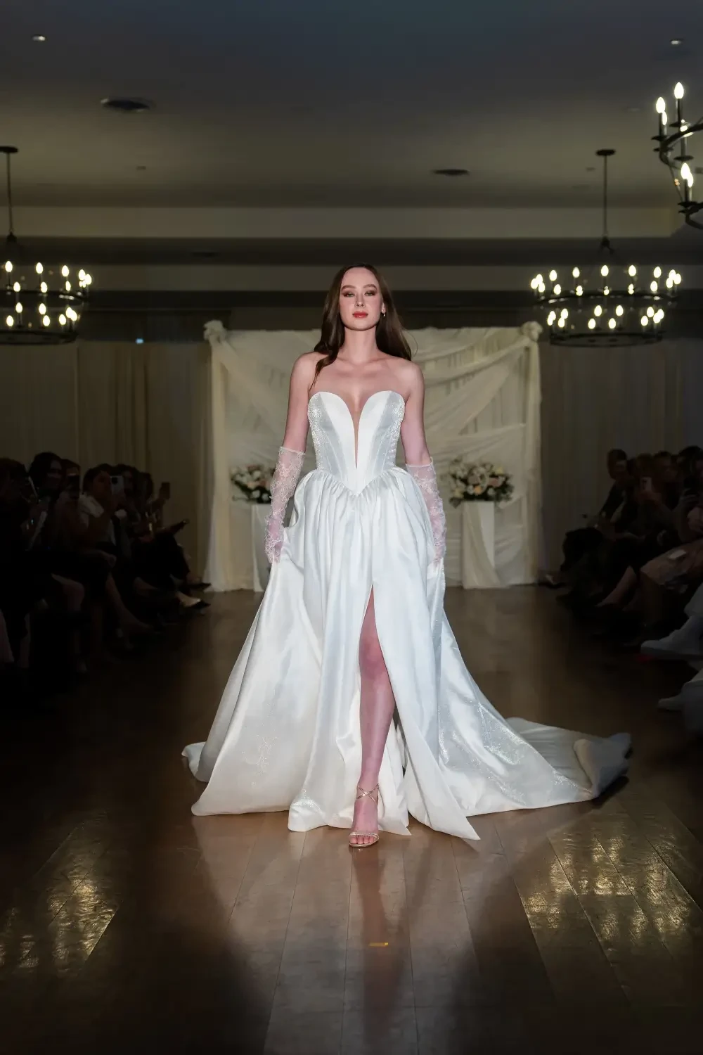 A model walks down a runway wearing a white wedding gown with a heart-shaped neckline and long gloves, surrounded by elegantly dressed attendees.