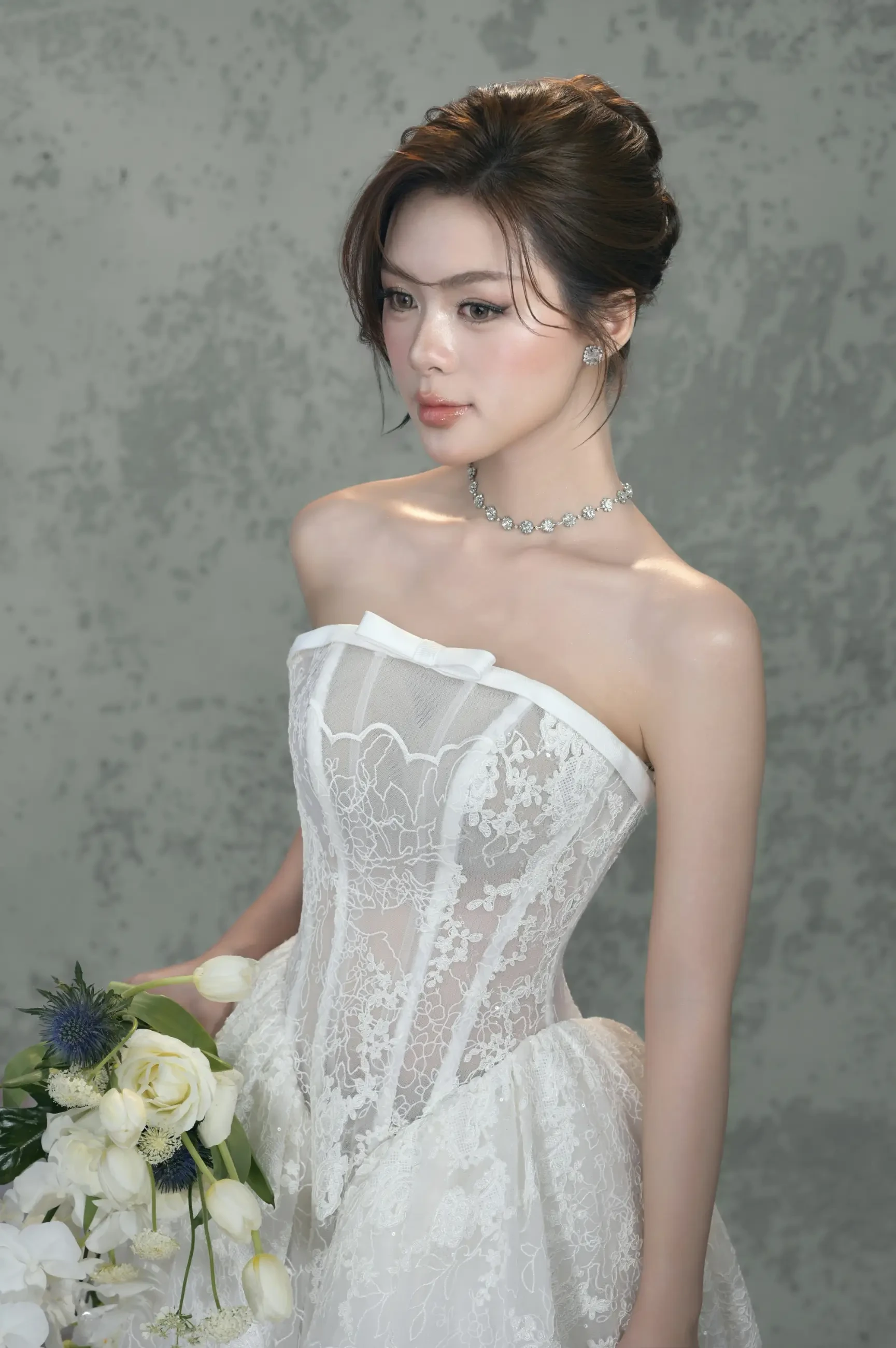 A woman in an elegant white lace wedding gown poses gracefully, holding a bouquet of white and blue flowers. She wears a sparkling necklace, exuding a serene and sophisticated ambiance against a textured gray background.