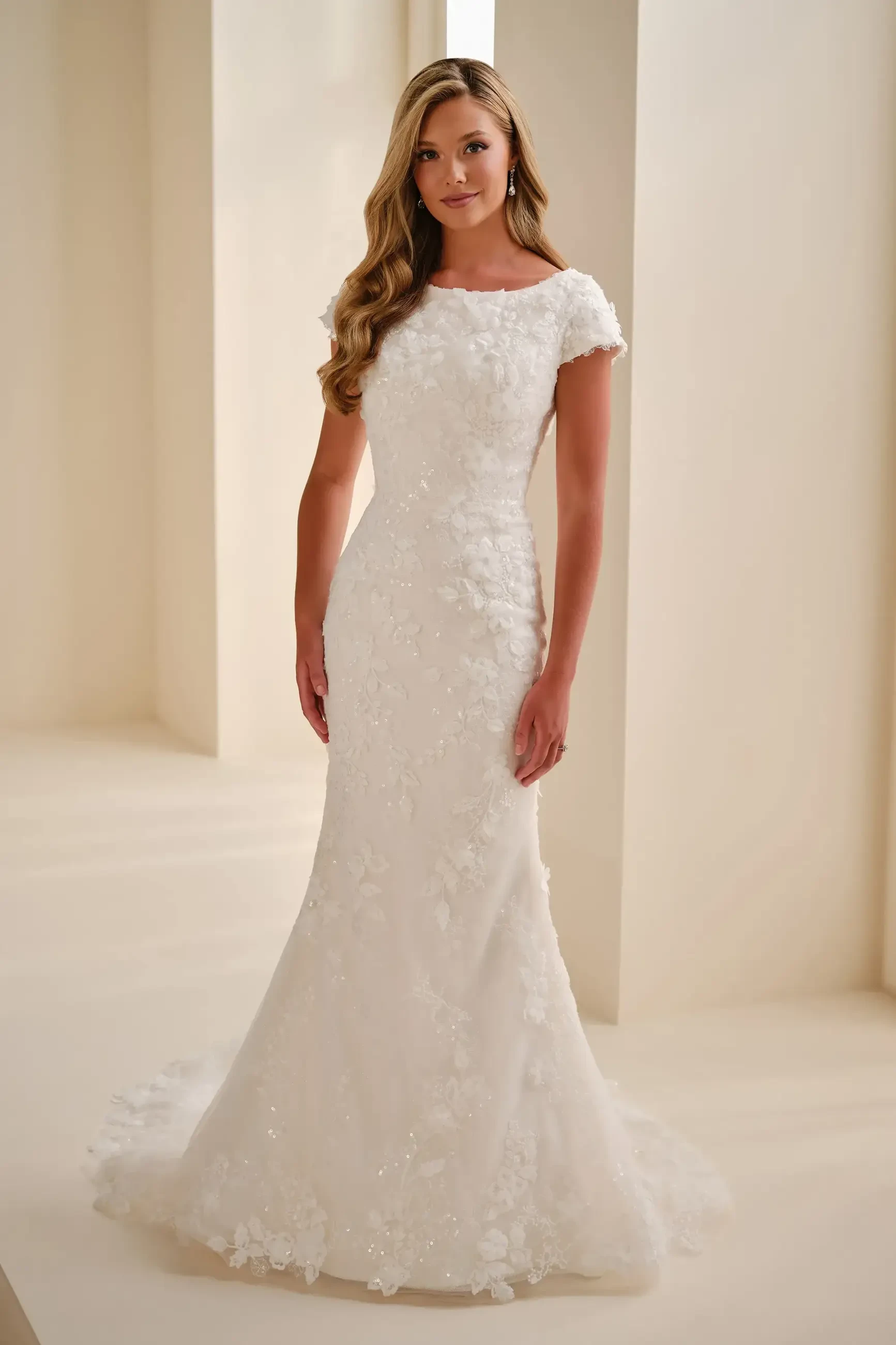 A woman in a white wedding dress with floral embellishments stands elegantly in a light, softly lit environment.
