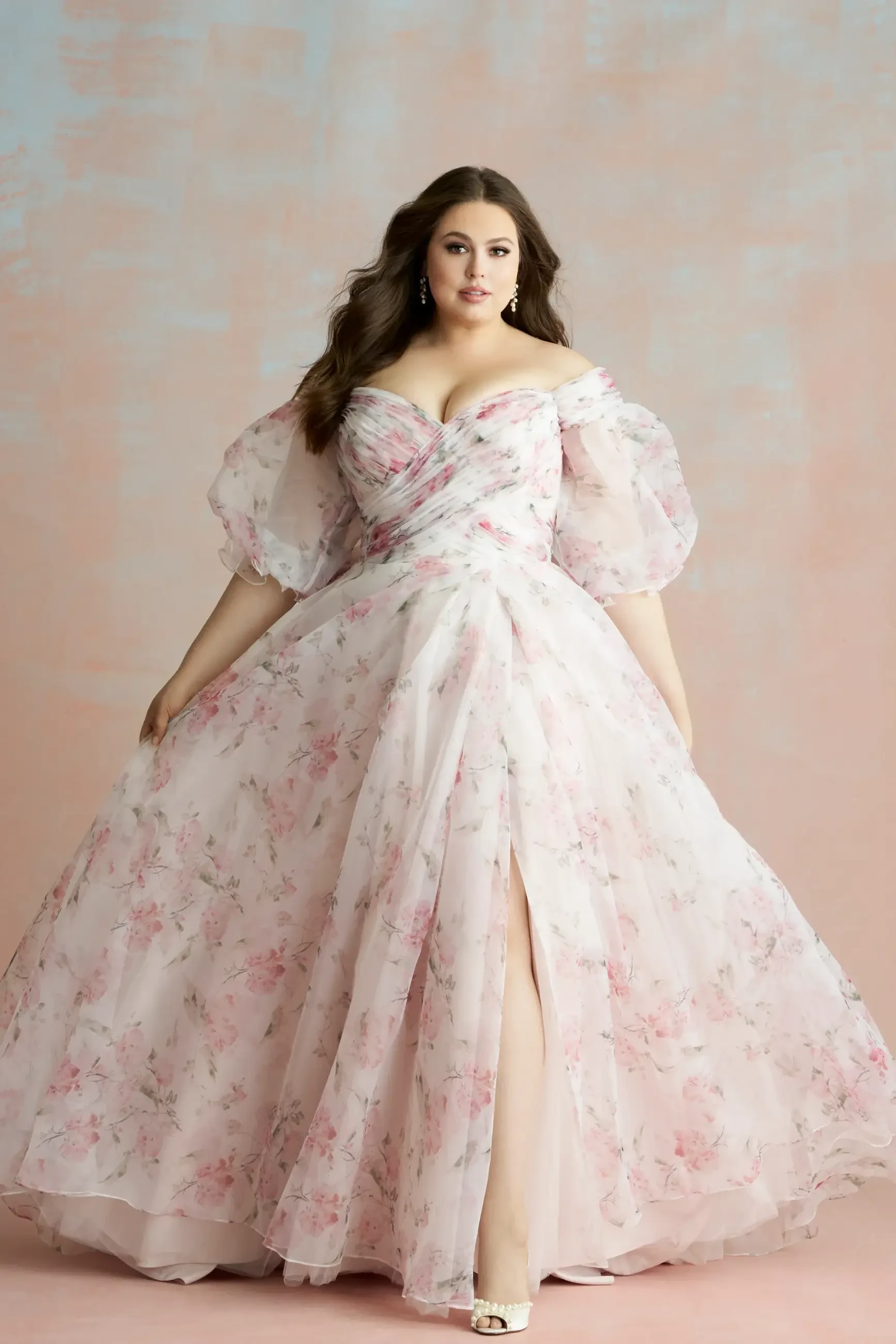 Model is wearing a soft A-line organza gown with a lavender and multicolored floral print, sweetheart neckline, off-the-shoulder sleeves, and a side slit.