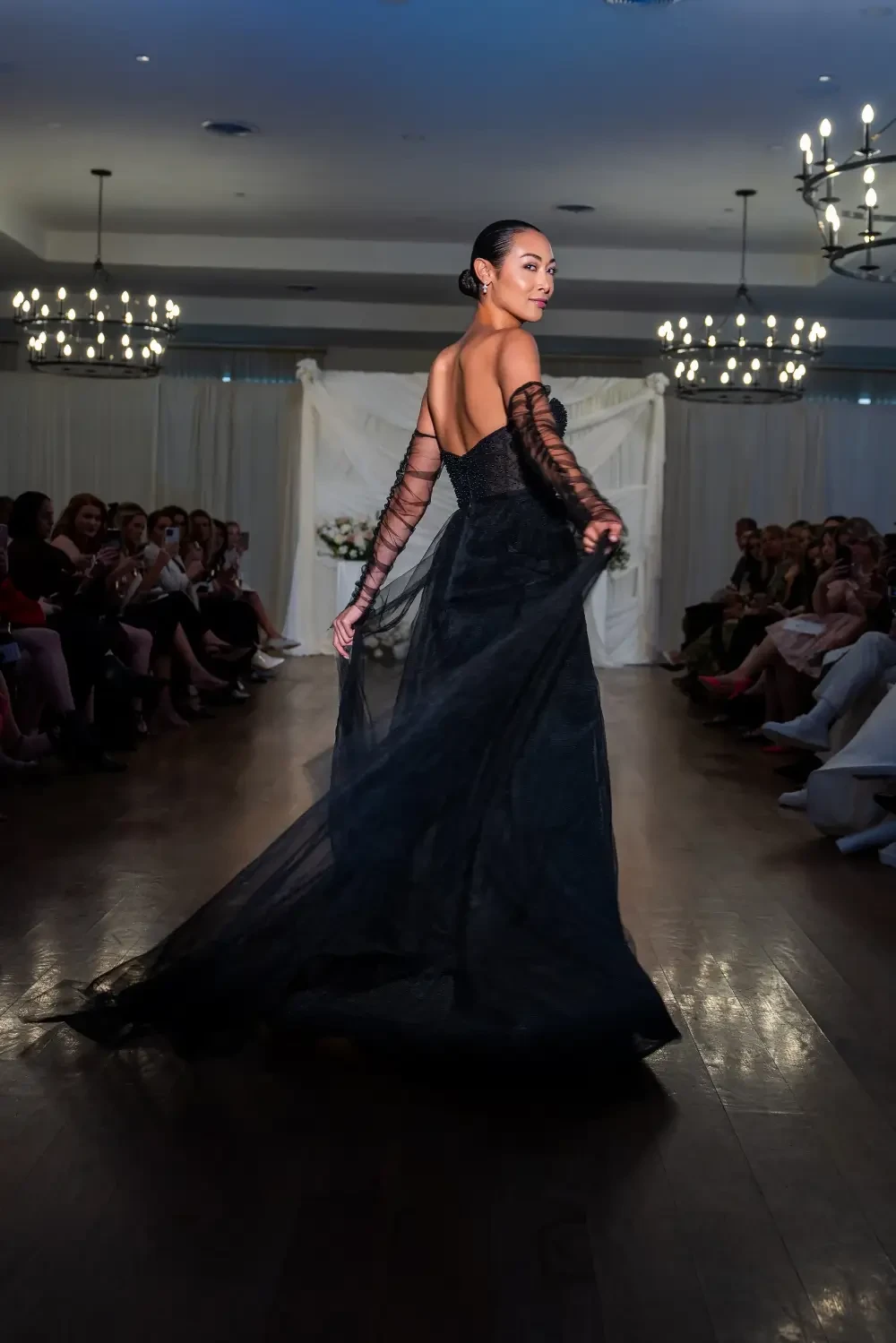 A model walks down a runway in a black gown, showcasing a flowing train, while an audience watches from the sides.