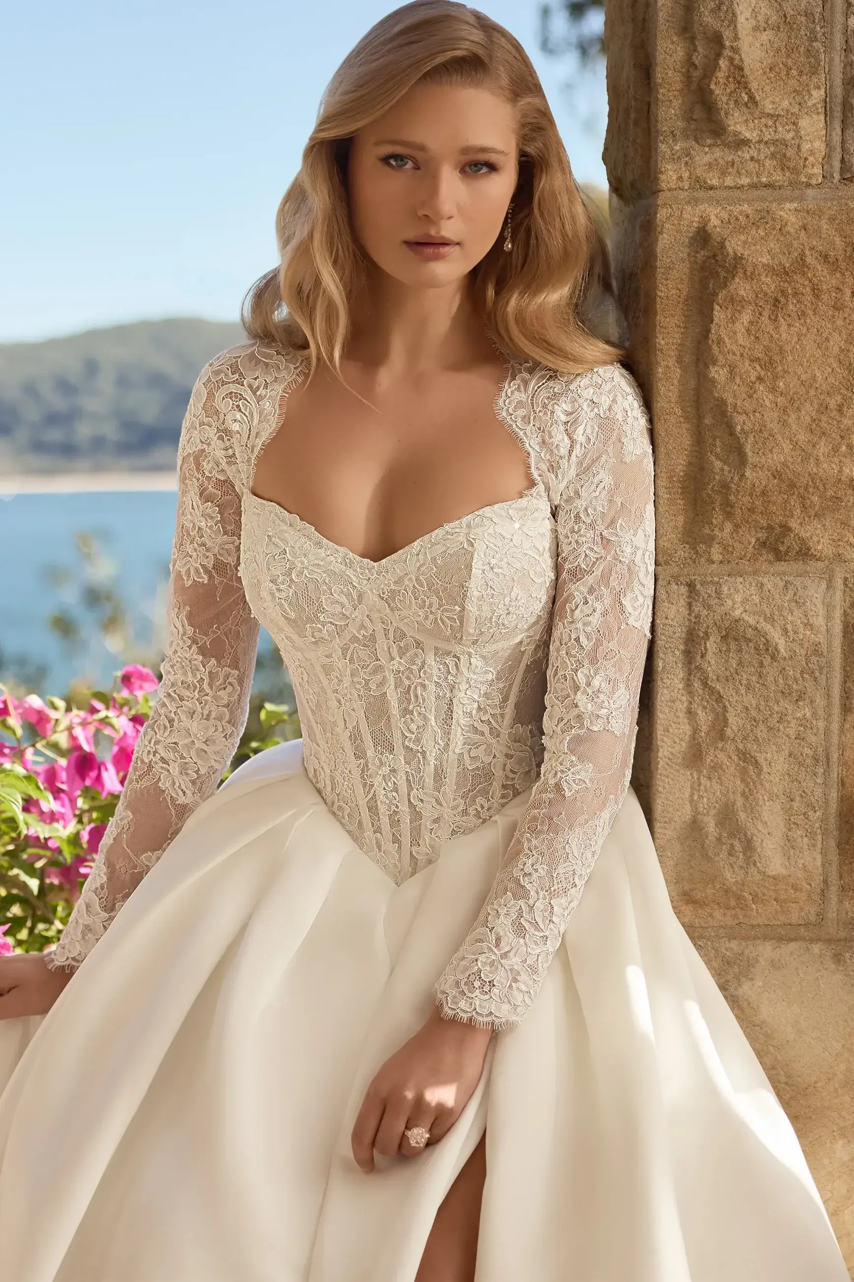 A woman in an elegant white wedding dress with long lace sleeves, posed next to a stone wall and surrounded by flowers, with a scenic view in the background.